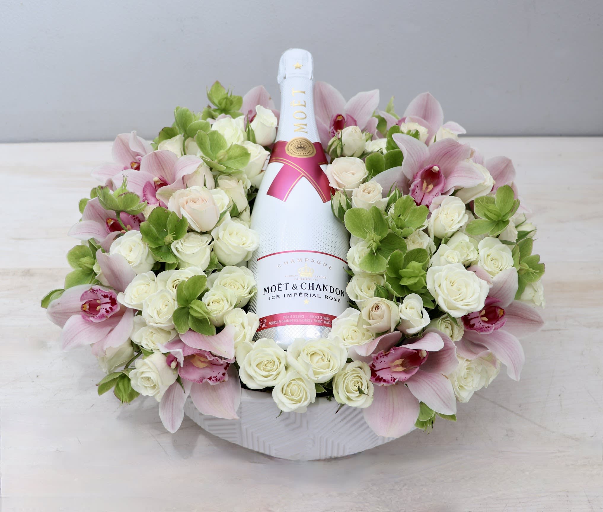 Bottle of champagne nestled in a low ring of white roses, green blooms, and pink orchids in a white bowl