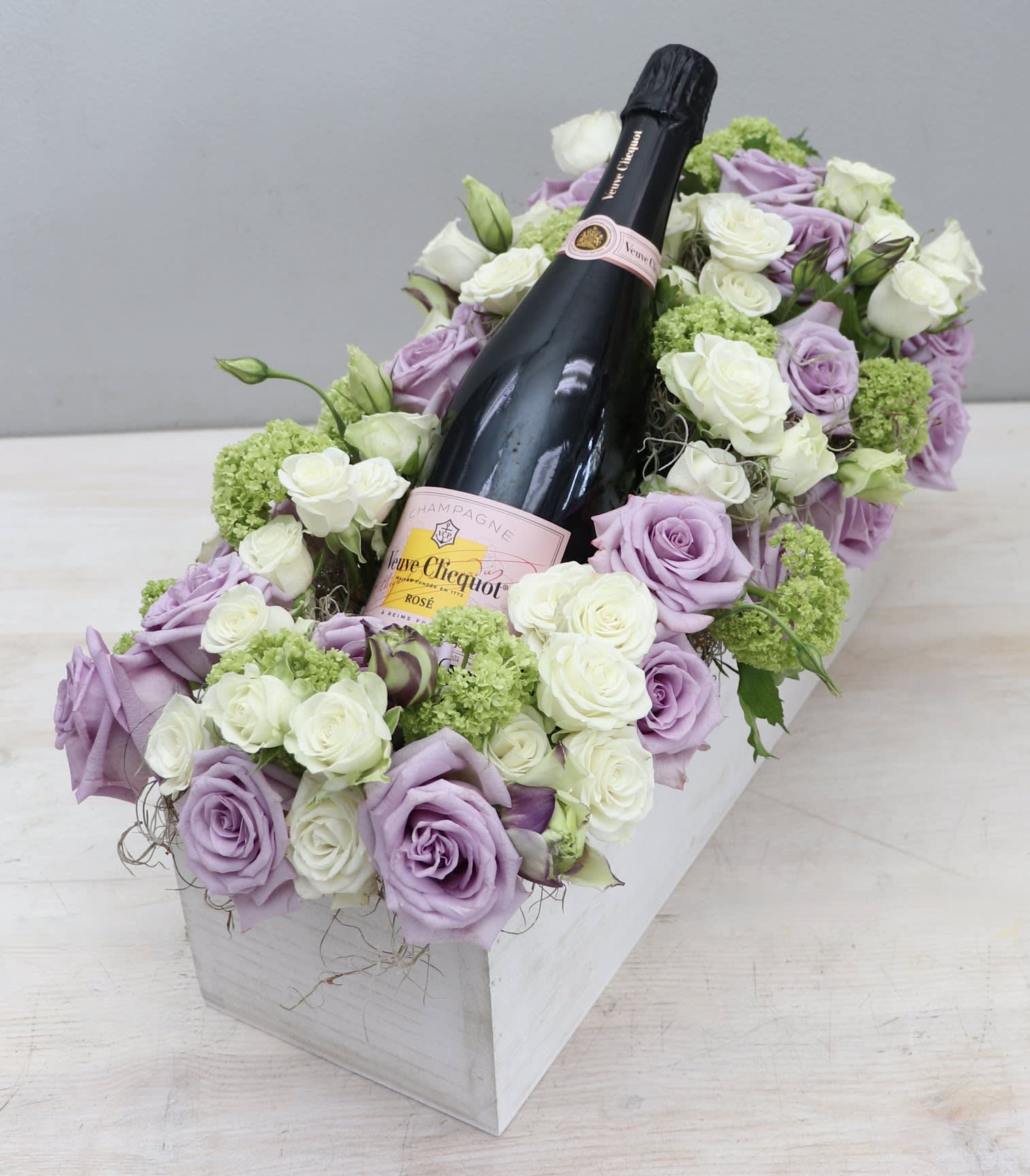 Rectangular box of lavender and white roses surrounding a rosé champagne bottle