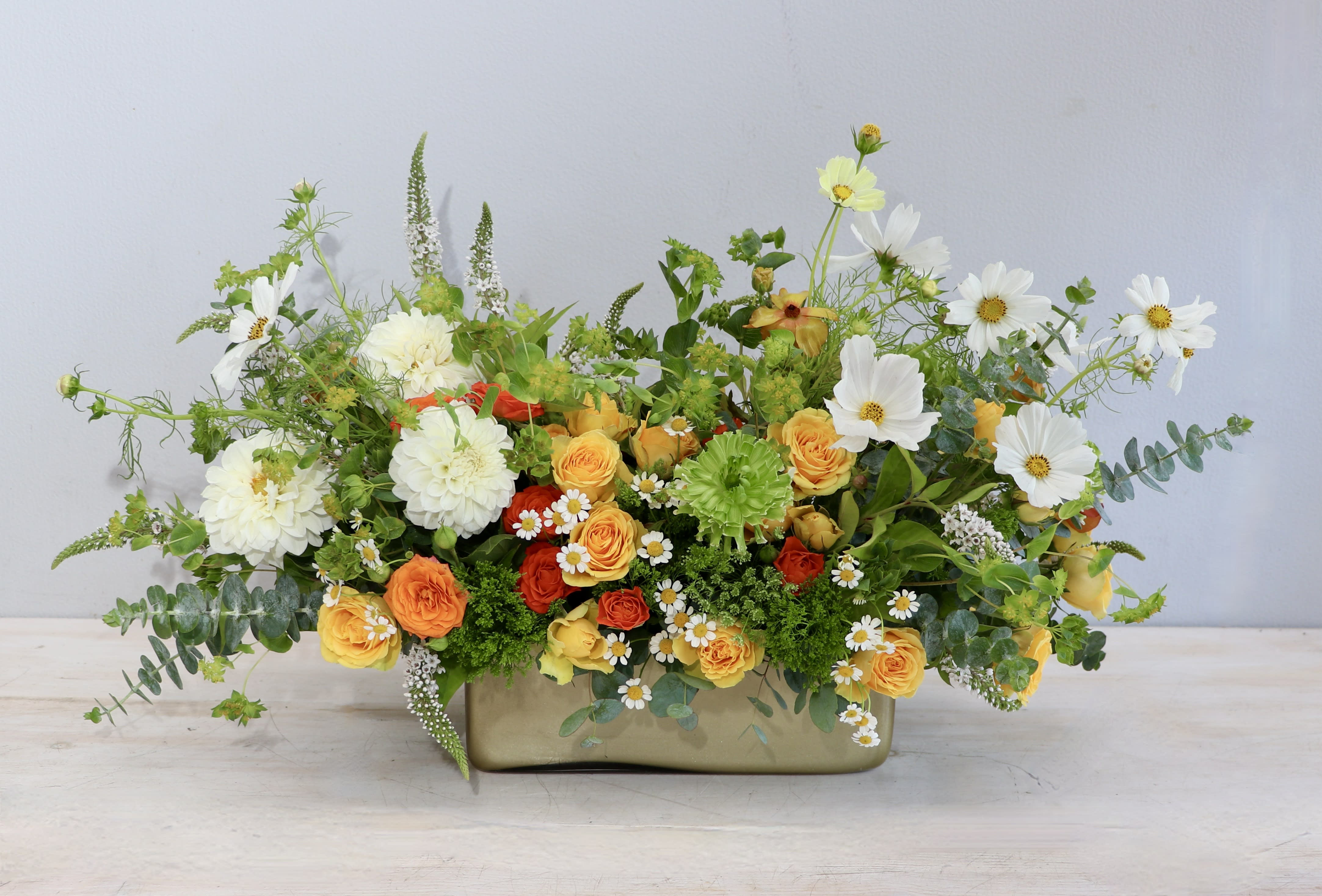 Low arrangement of yellow and orange roses with white blooms in a rectangular gold vase