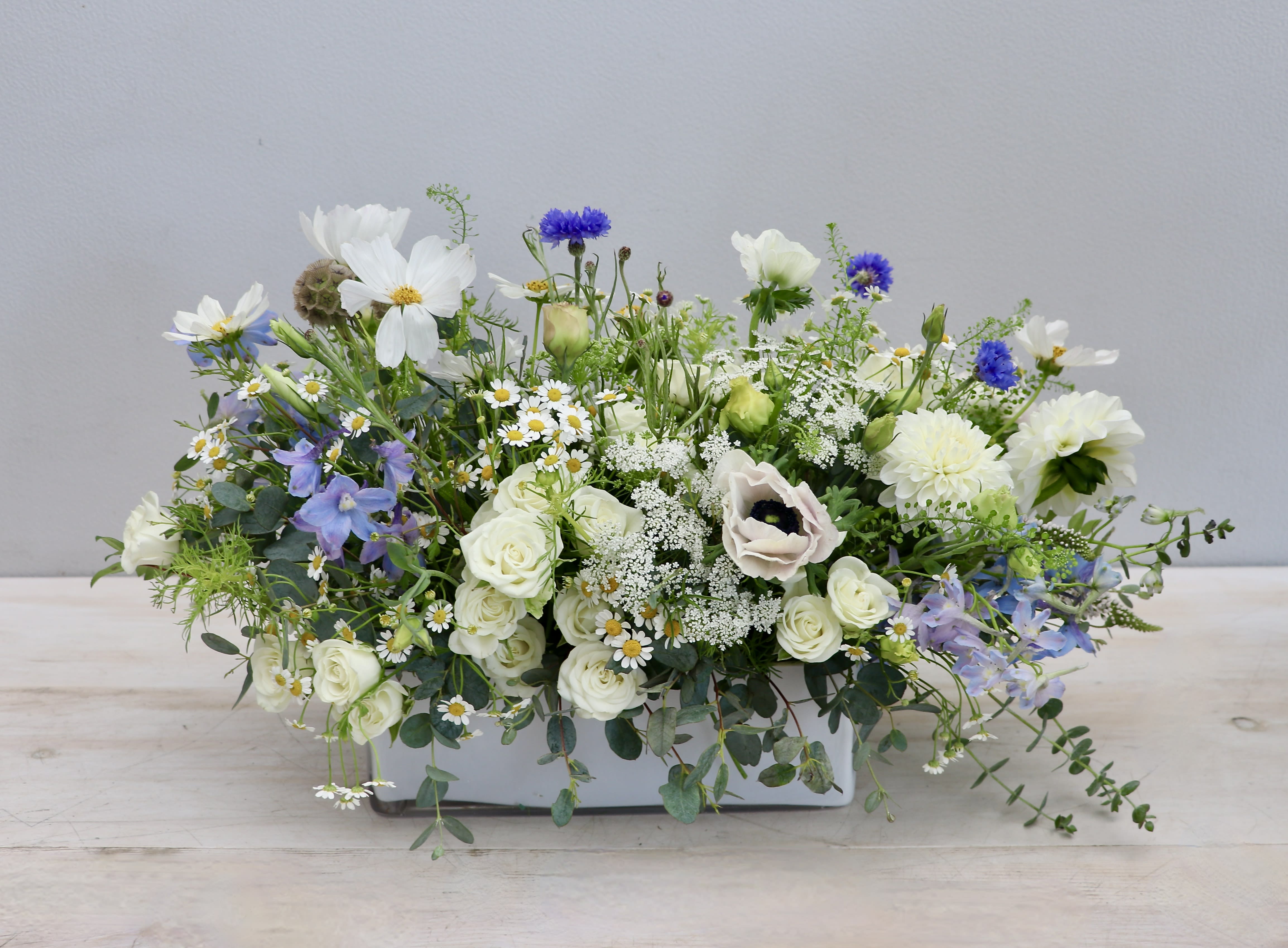 Low white ceramic centerpiece with white, blue, and purple flowers in a loose garden style