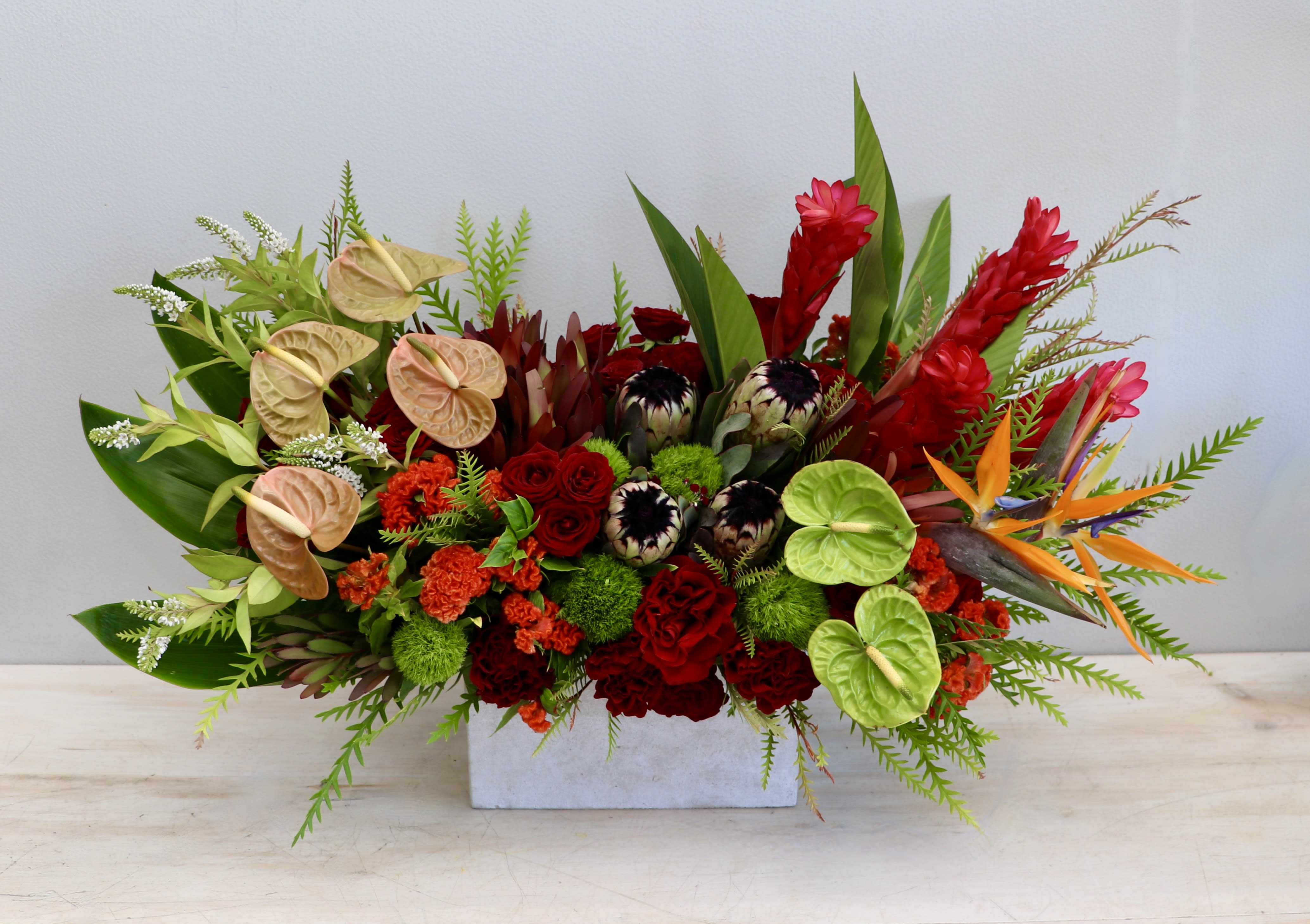 Long centerpiece of red and orange tropical flowers with green and peach anthuriums in a white rectangular vase