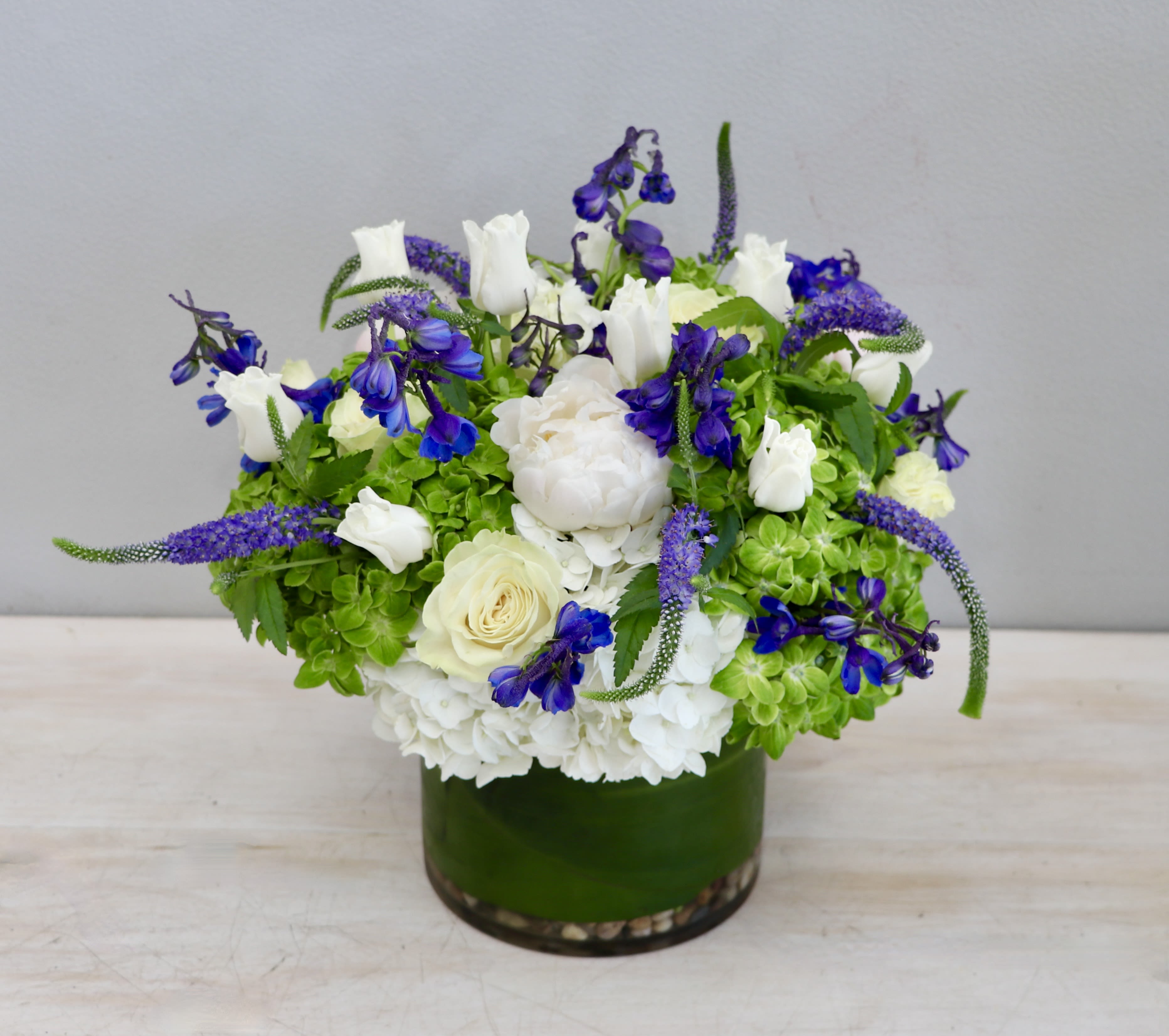 Low round arrangement of white and blue flowers with green accents in a glass vase wrapped with a green leaf.