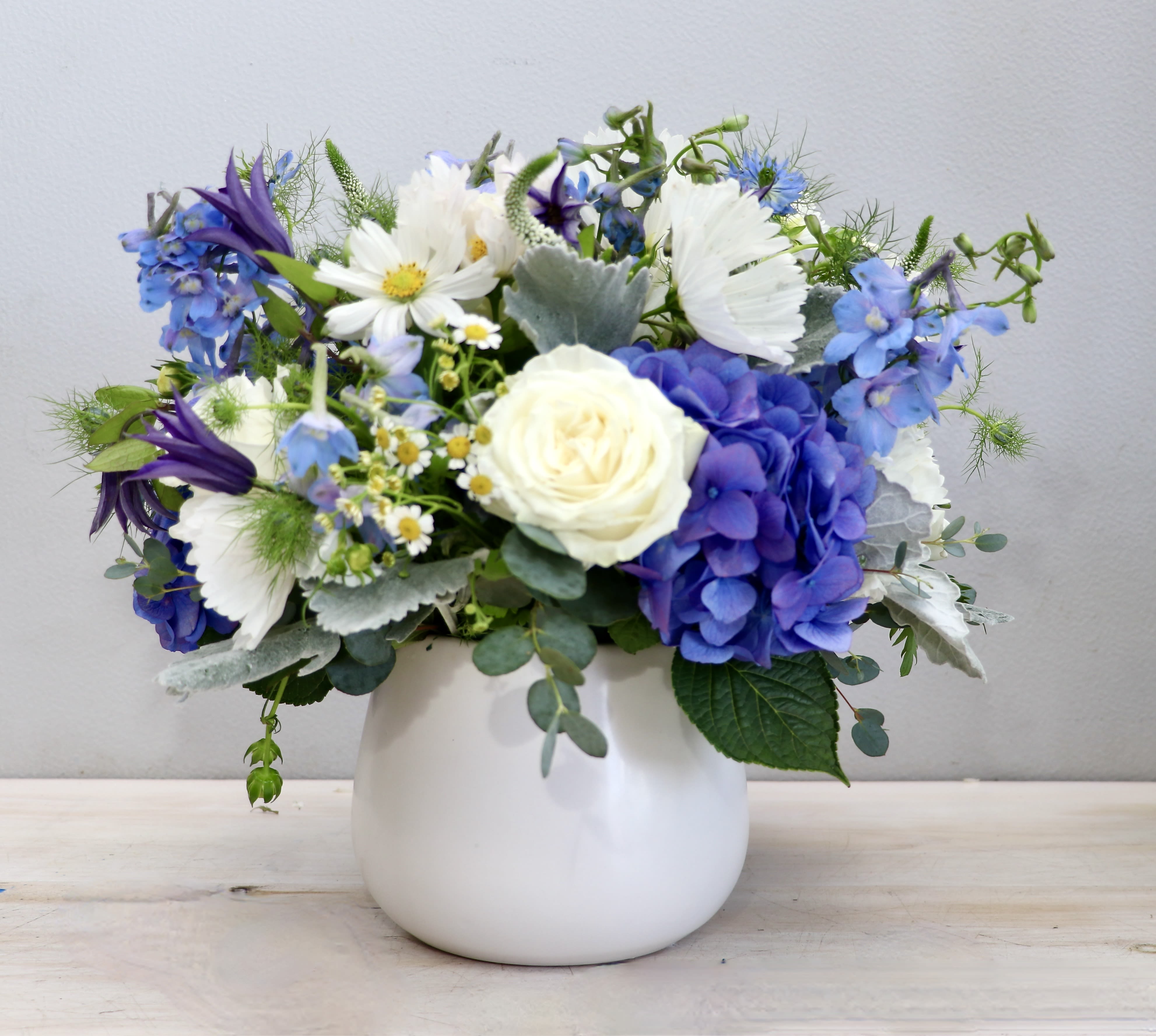 Short blue and white flower arrangement with hydrangeas and roses in a round white vase