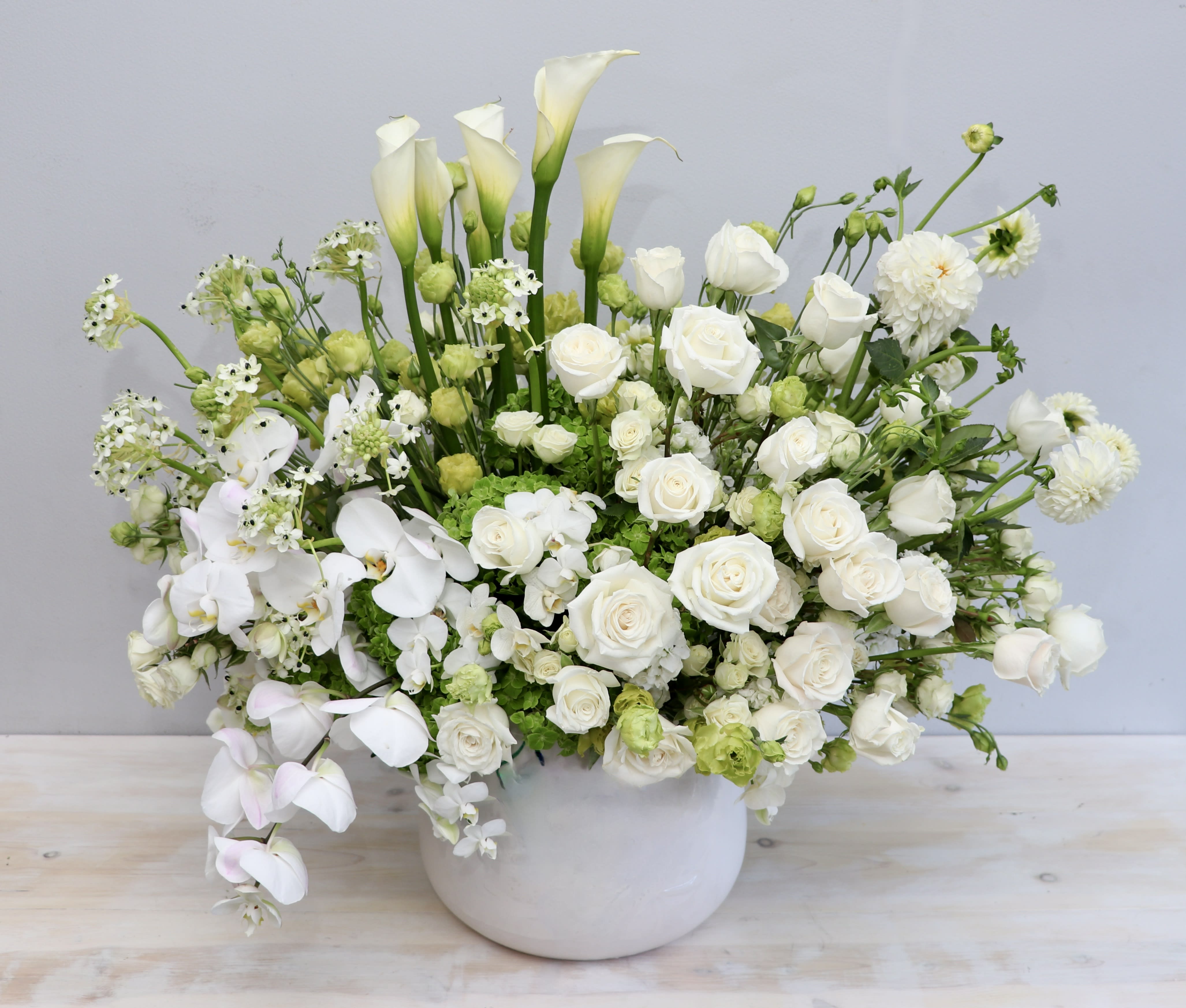 Large white and green arrangement of roses, calla lilies, orchids, and other blooms in a white ceramic bowl