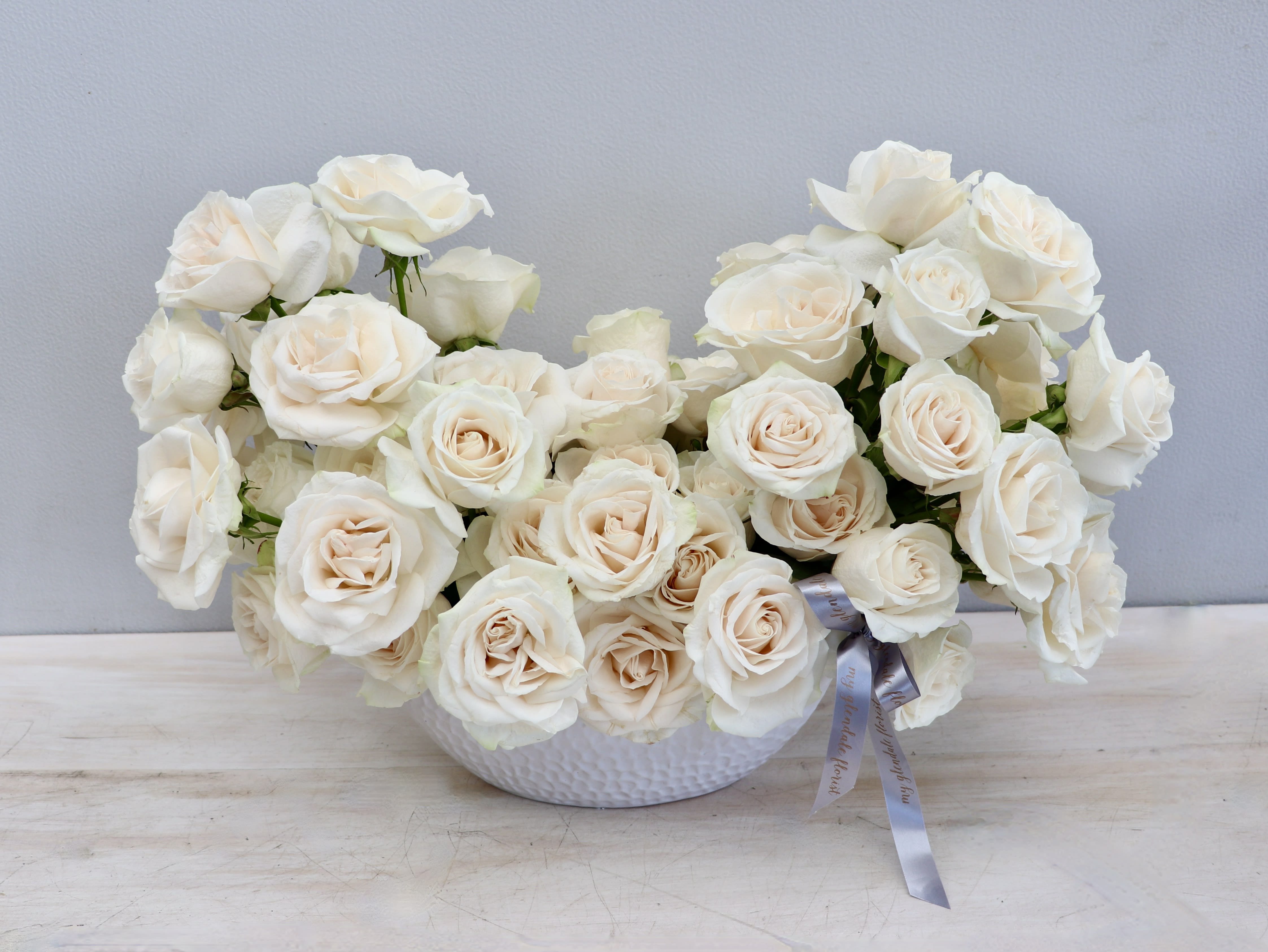 Low white bowl of clustered ivory roses with a gray ribbon accent on a light wood surface
