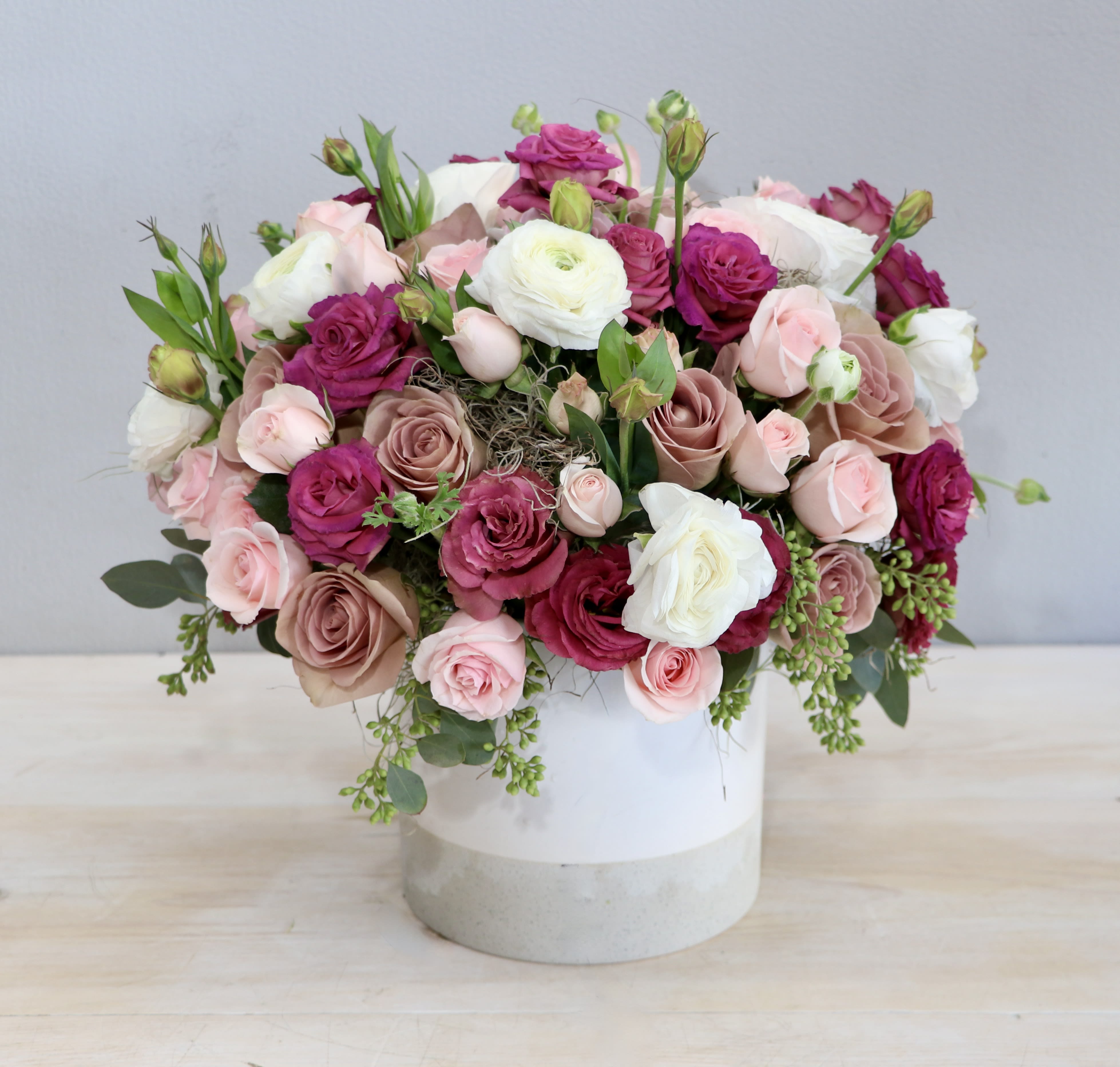 Low arrangement of pink, mauve, and white roses and ranunculus in a white cylinder vase
