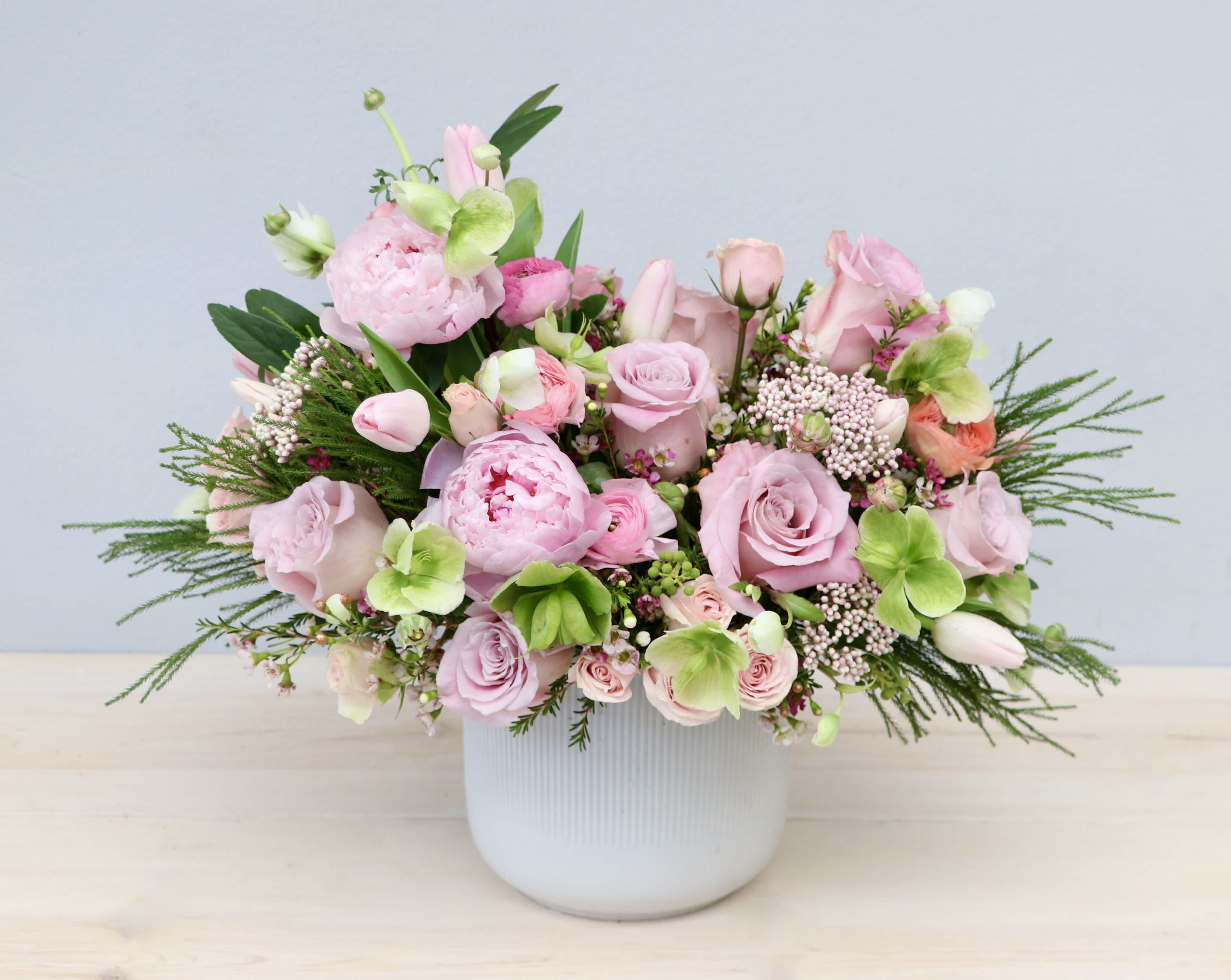 Low arrangement of soft pink roses, peonies, and tulips with light green blooms in a white ceramic pot