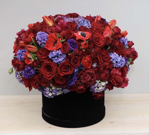 Low dome of red roses and purple blooms arranged in a black hatbox-style container