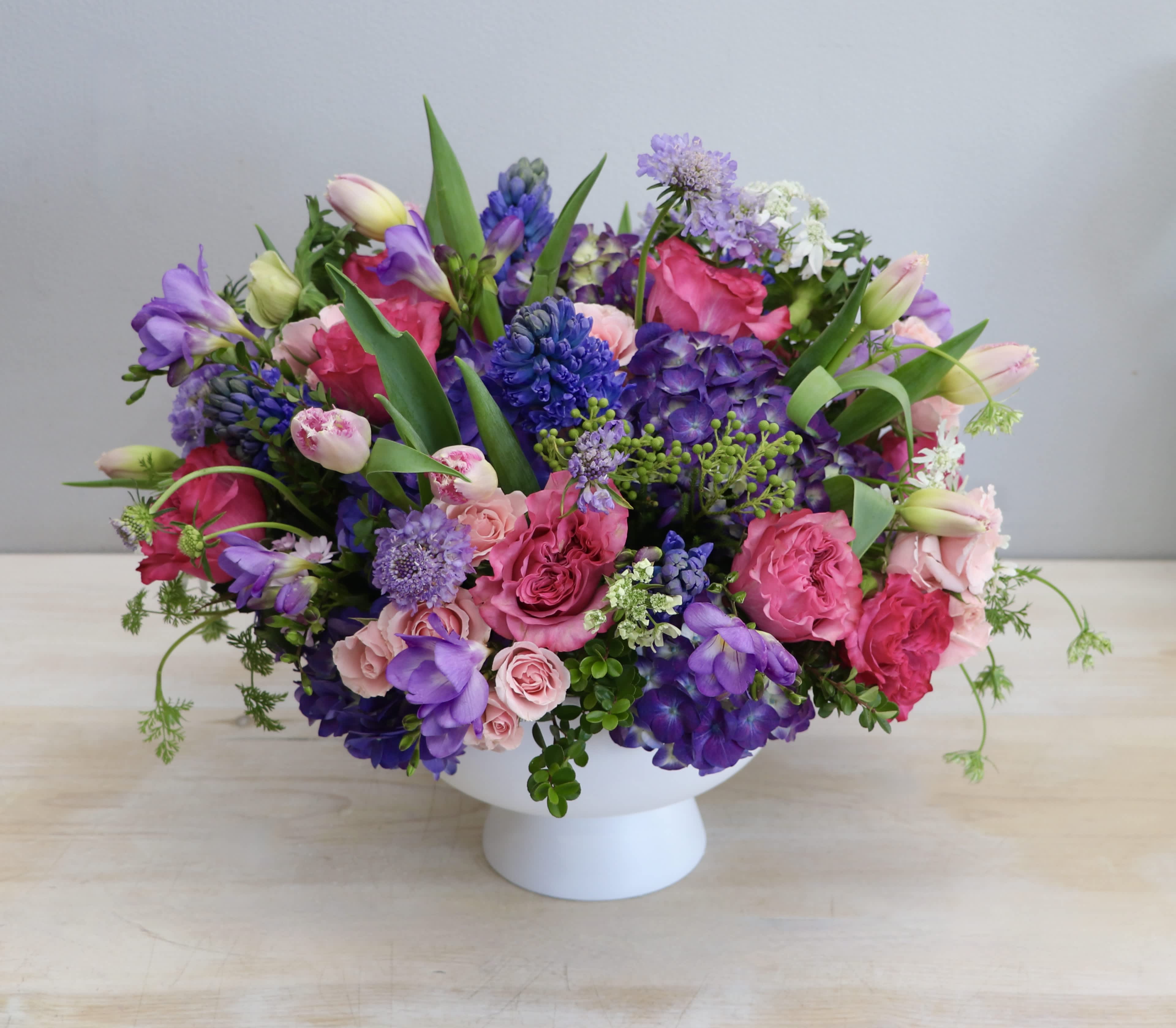 Low arrangement of pink roses, tulips, and purple blooms in a white pedestal bowl