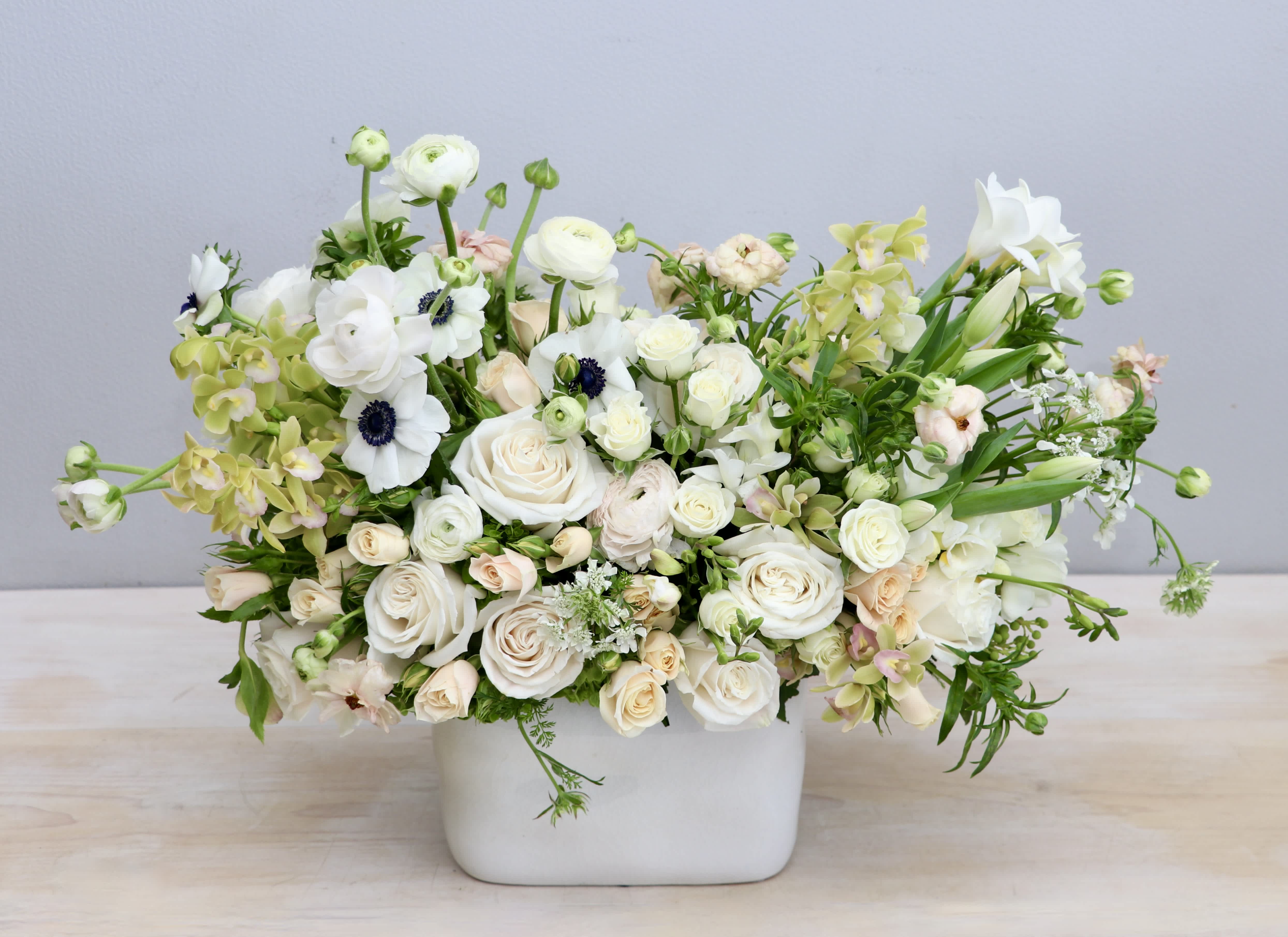 Low white and cream flower arrangement with roses, ranunculus, anemones and orchids in a rectangular white vase.