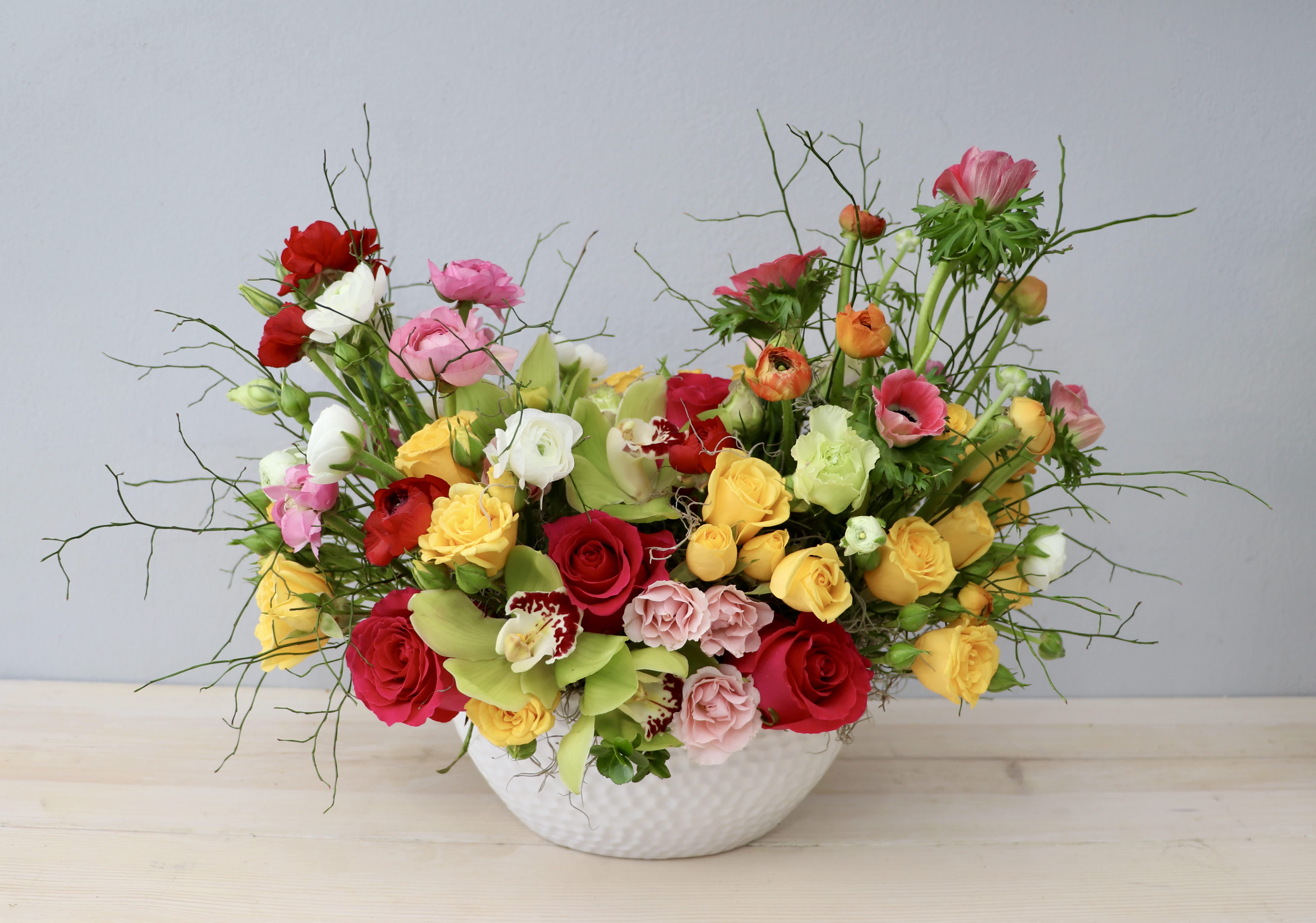 Low white bowl of mixed yellow, red, and pink roses with ranunculus and orchids in a loose, colorful arrangement