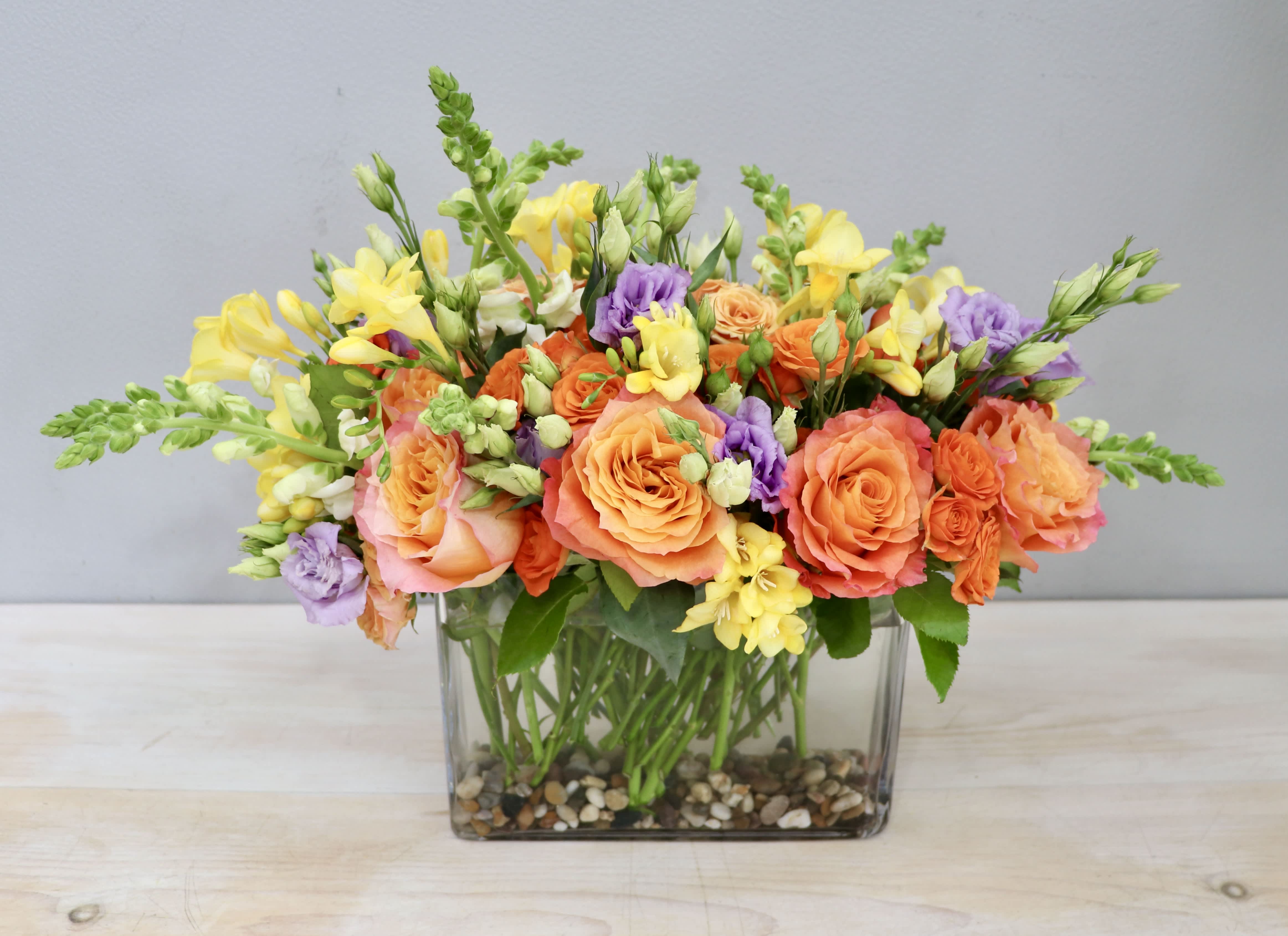 Low rectangular arrangement of orange roses, yellow flowers, and purple blooms in a glass vase with pebbles