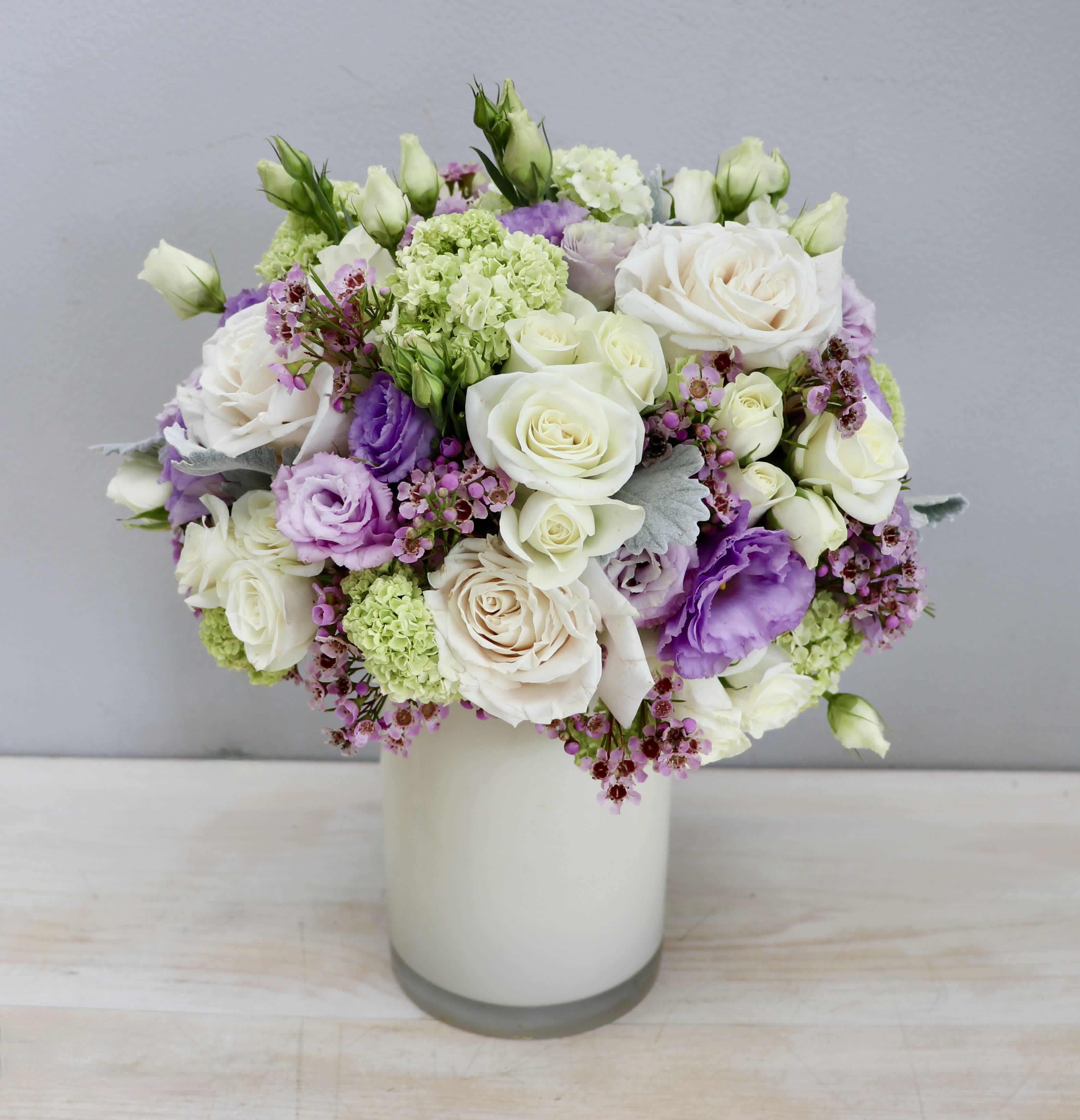 Compact arrangement of white roses and purple lisianthus in a white cylinder vase