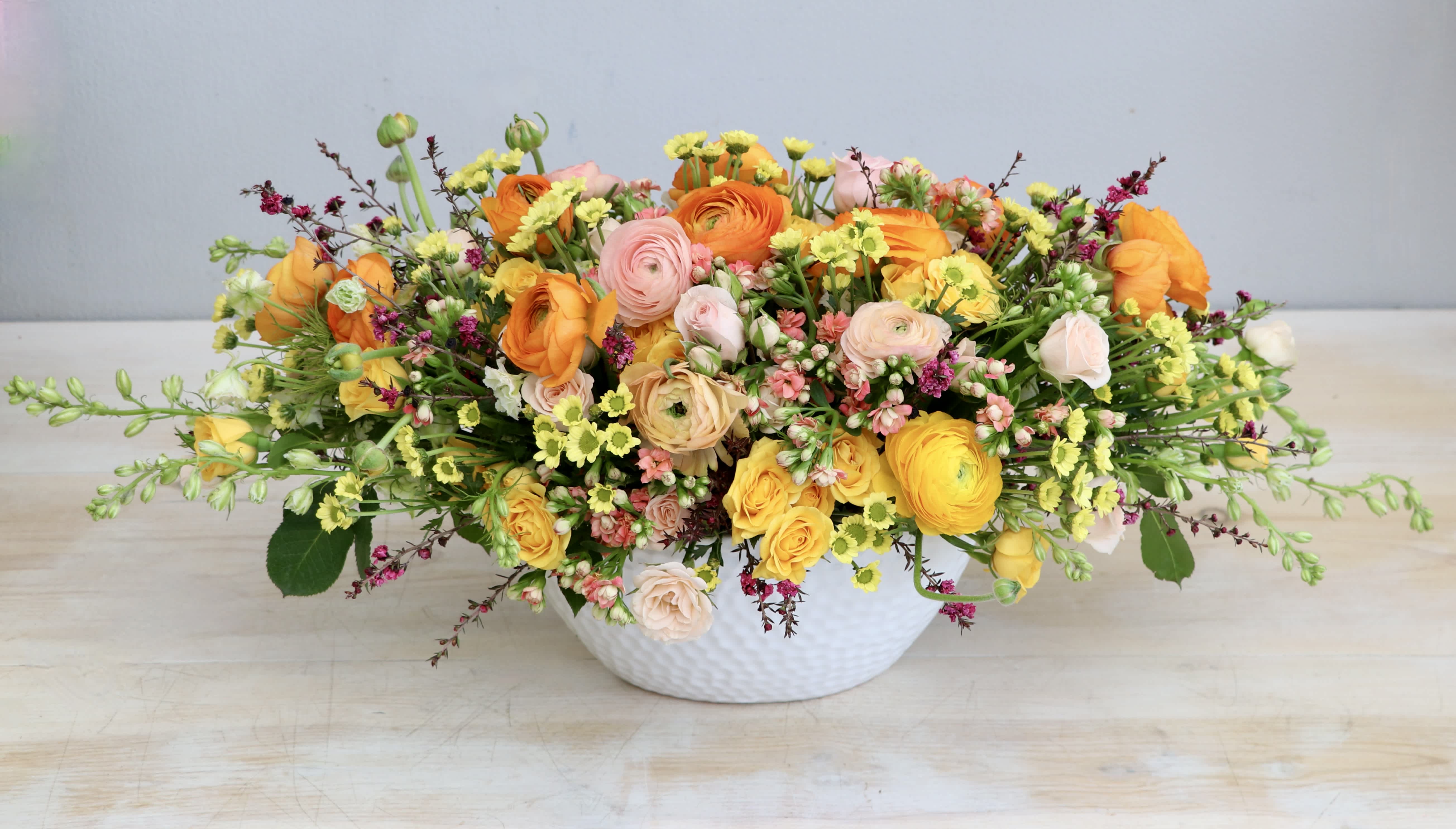 Low centerpiece of yellow, orange, and blush flowers arranged in a white ceramic bowl
