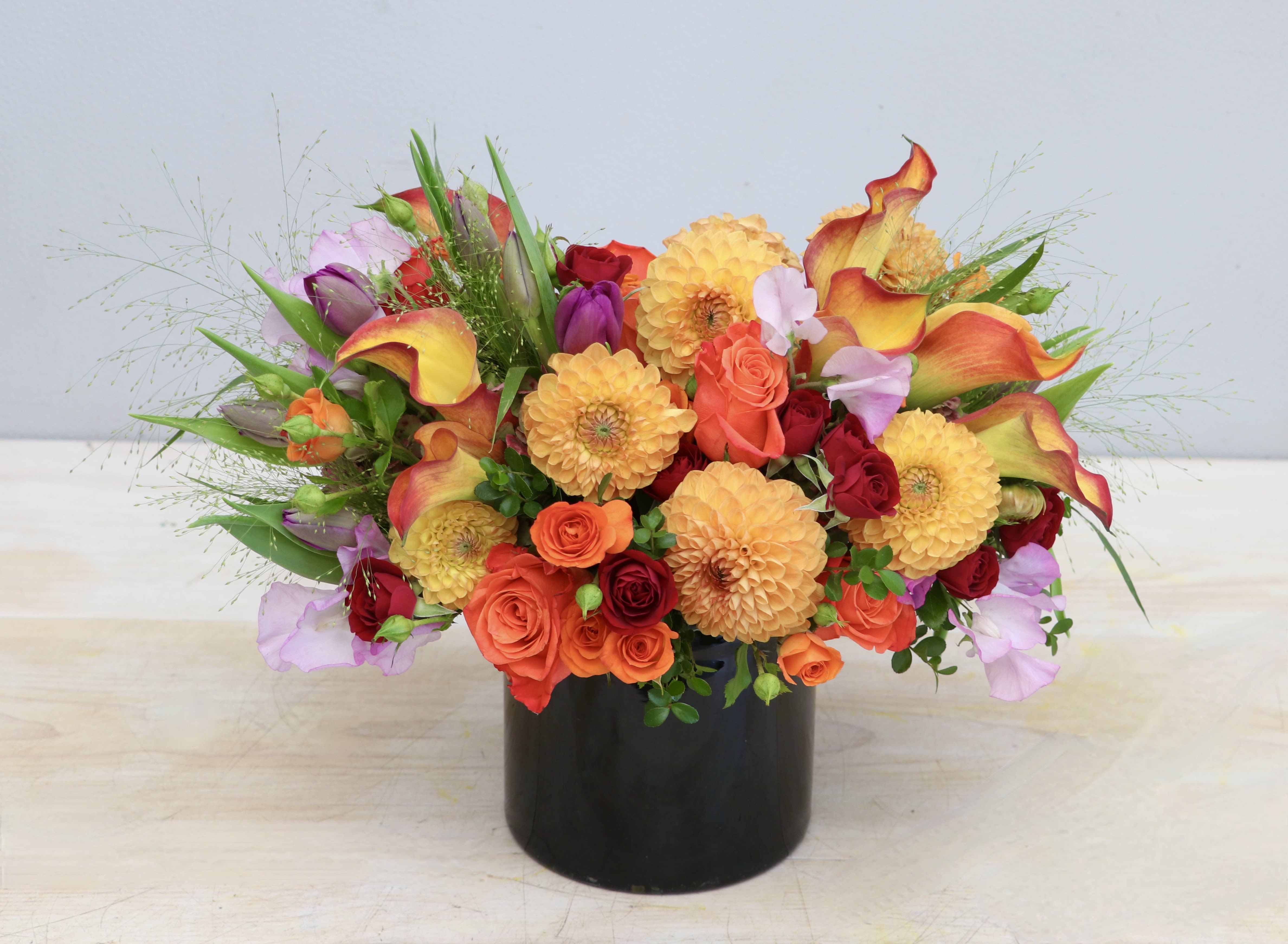 Bright mixed arrangement of orange dahlias, roses, and calla lilies in a black cylinder vase