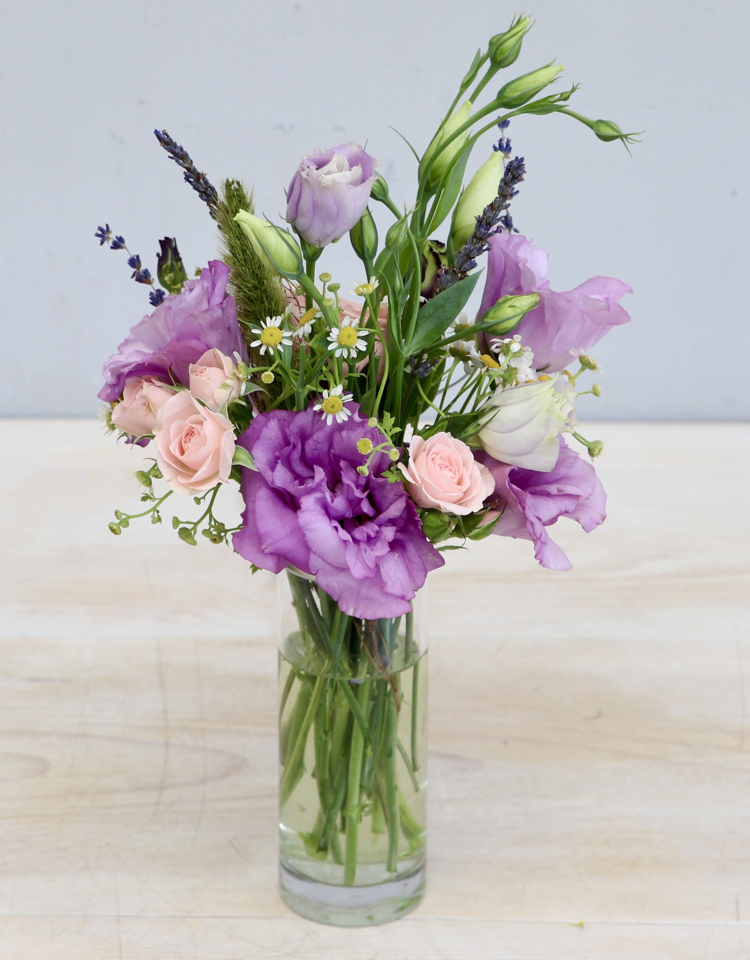 Tall glass vase with purple lisianthus, pink spray roses, and small white daisies in a loose arrangement