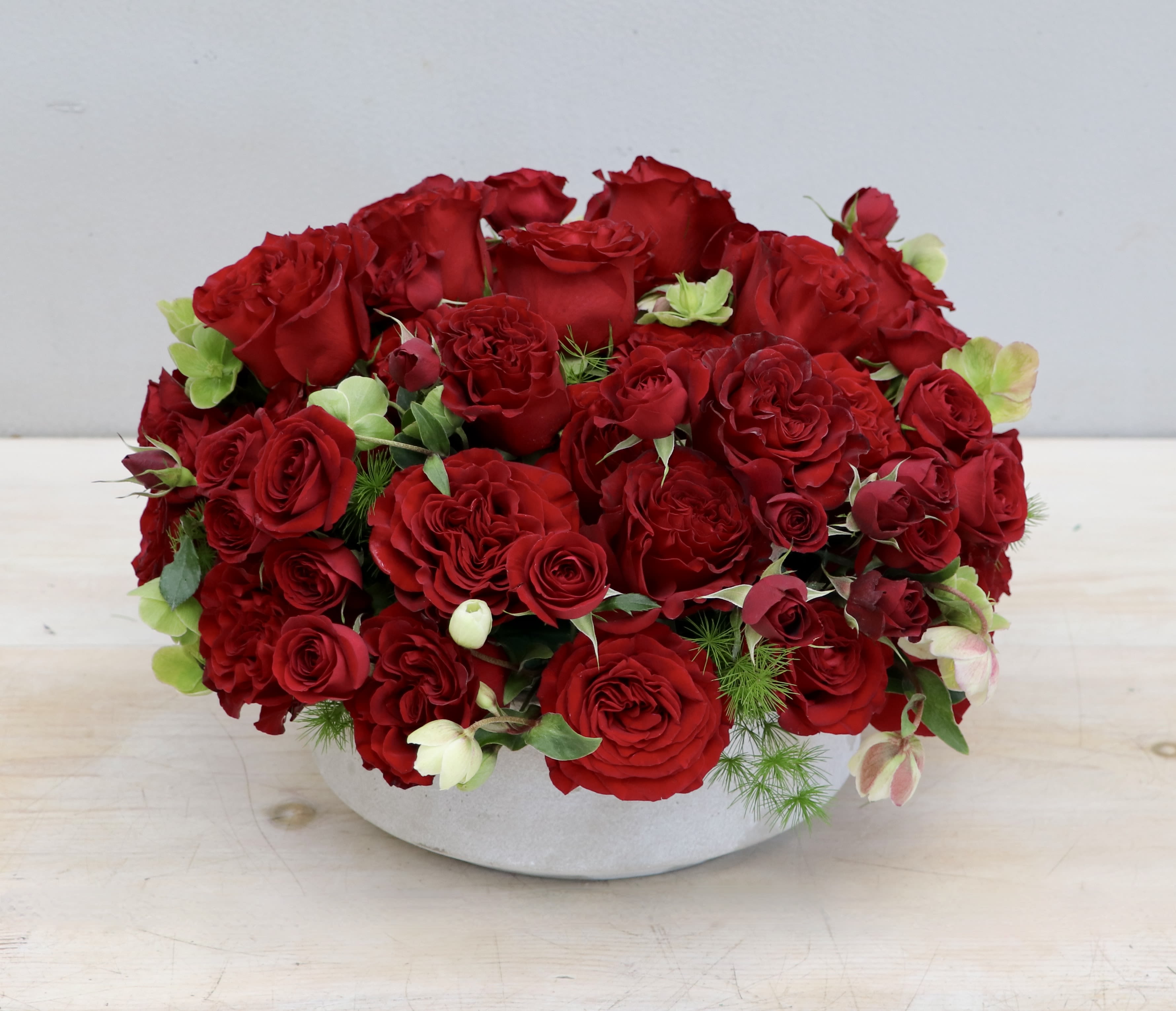Low round arrangement of red roses in a white ceramic bowl with small green floral accents.