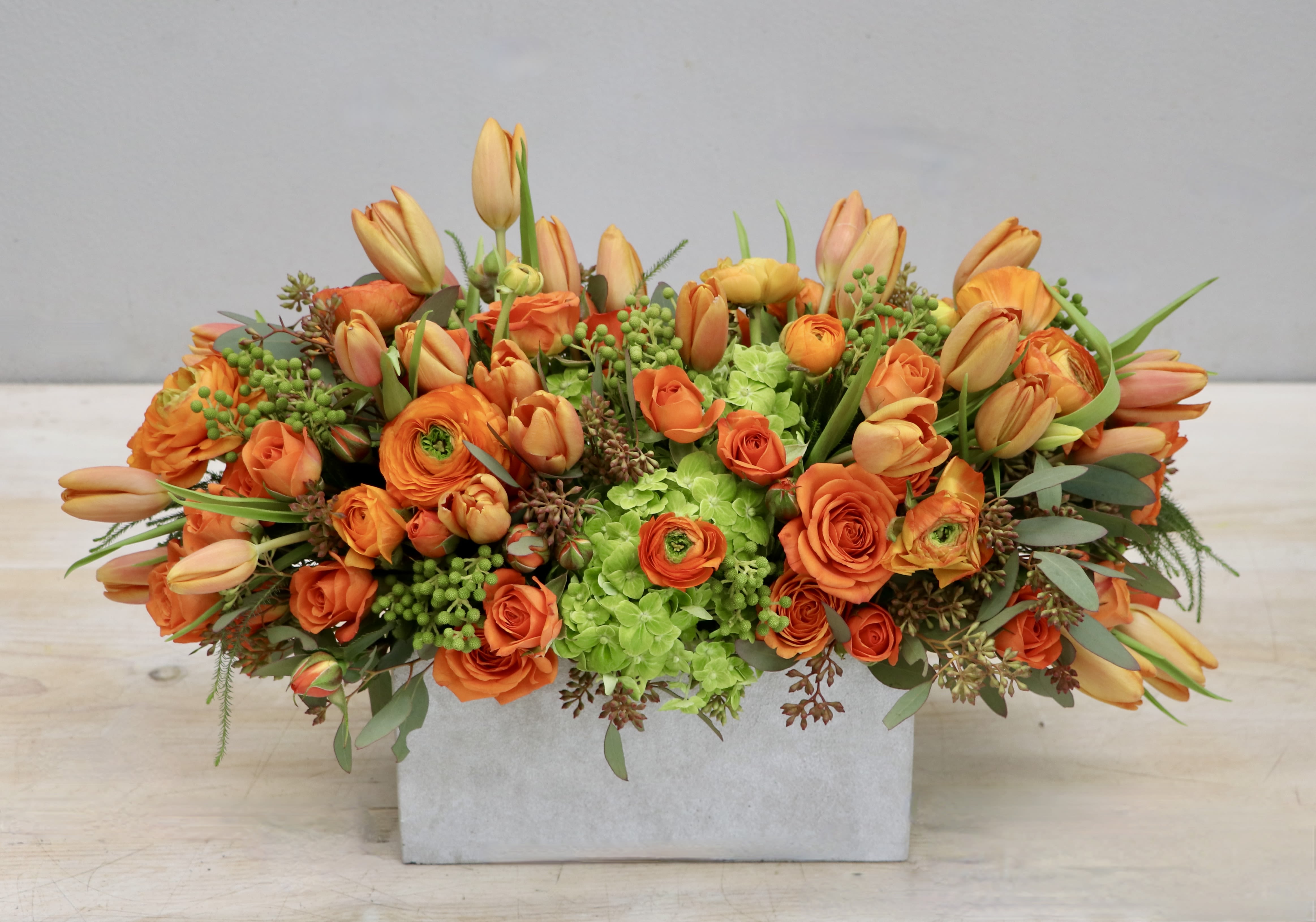 Low rectangular arrangement of bright orange roses, tulips, and ranunculus with green hydrangeas