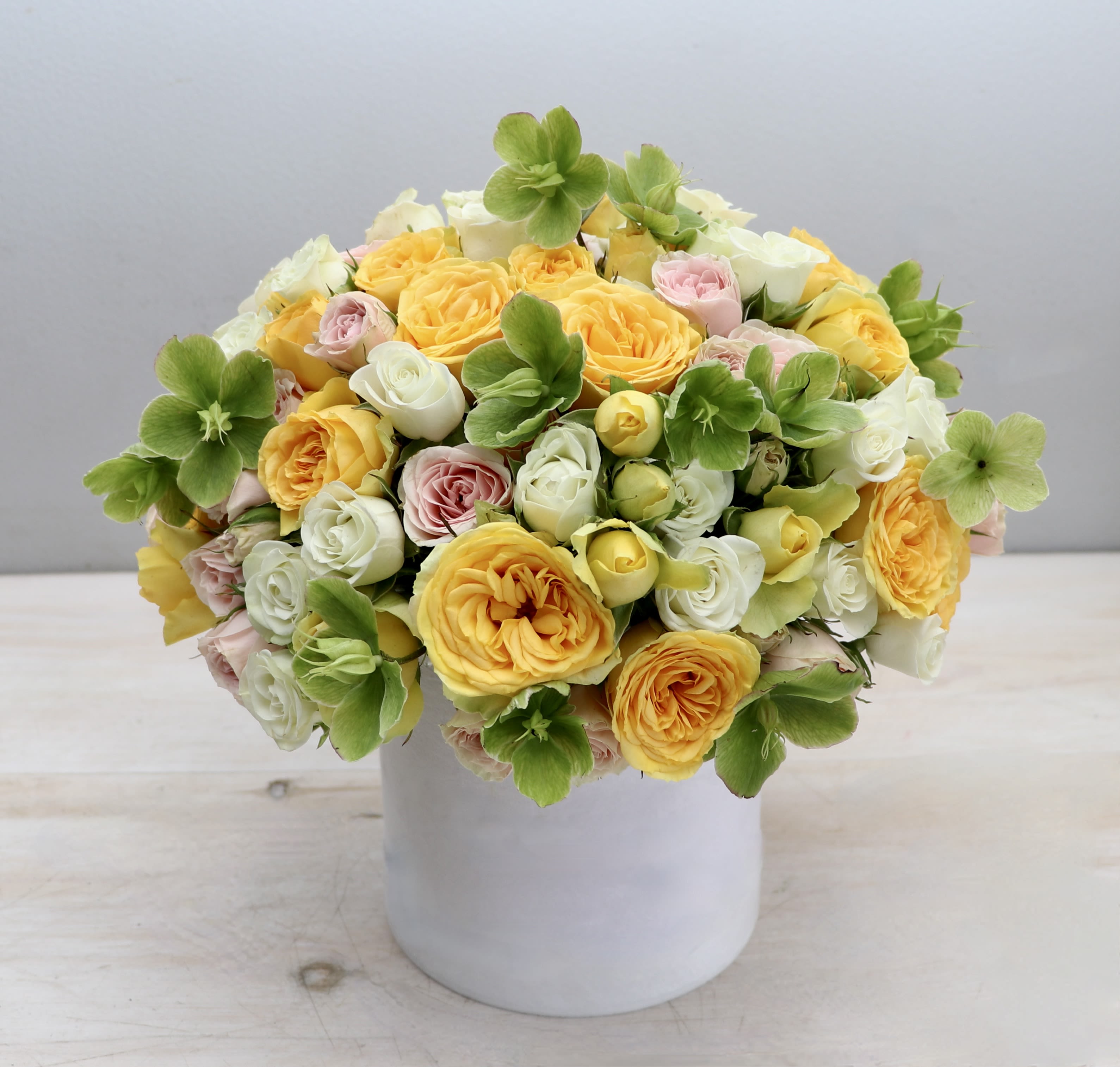 Low arrangement of yellow and pastel roses with green blooms in a white ceramic cylinder vase