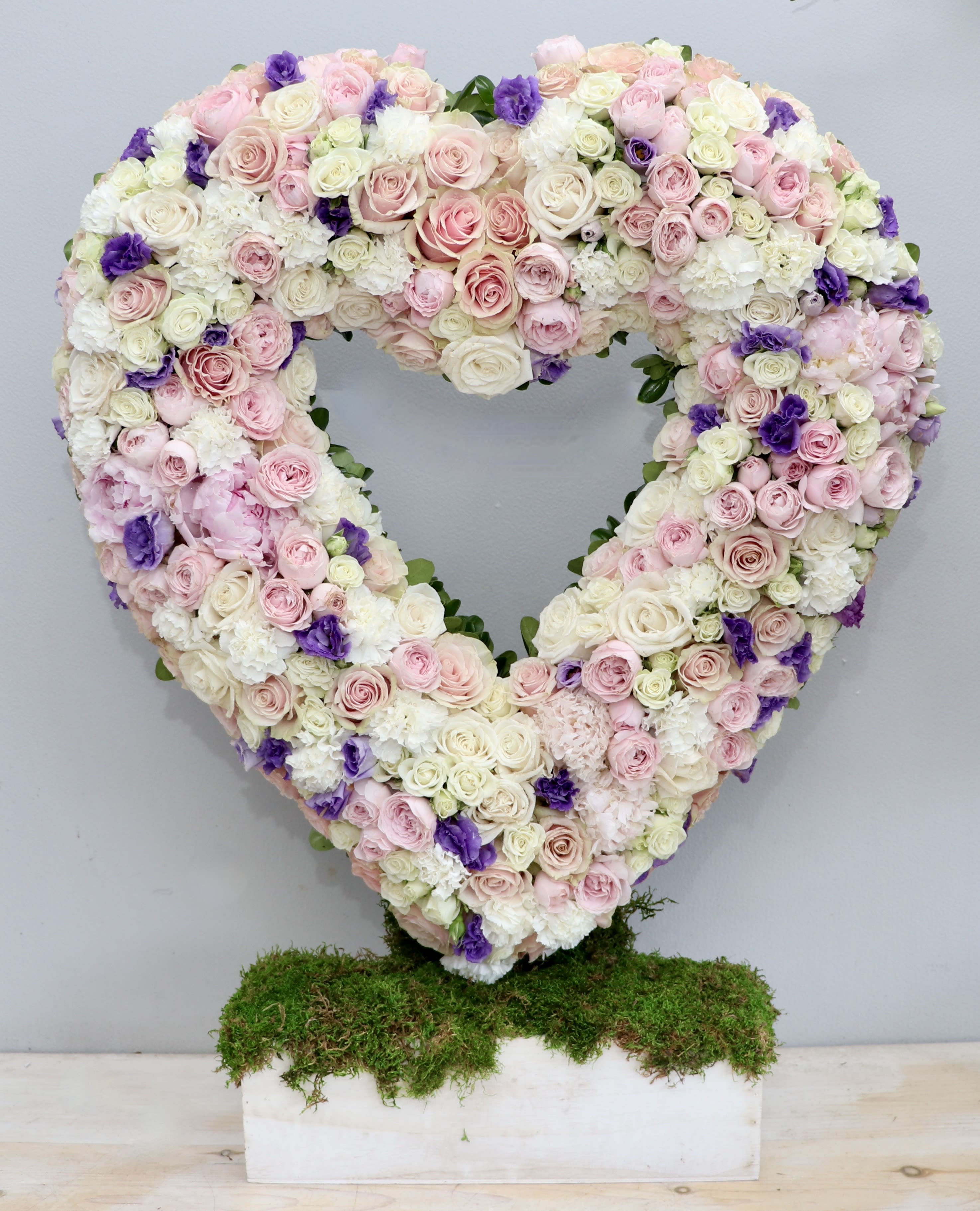 Heart-shaped standing arrangement of pink and white roses with purple accents on a moss-covered base.