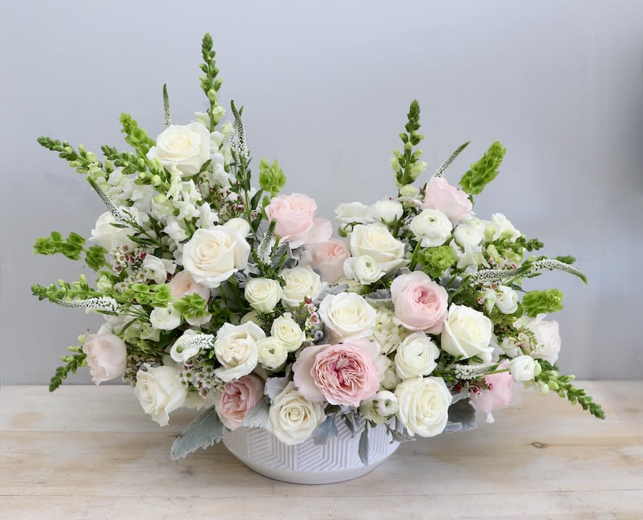 Low white bowl of white and blush roses with green accents in a wide, airy arrangement