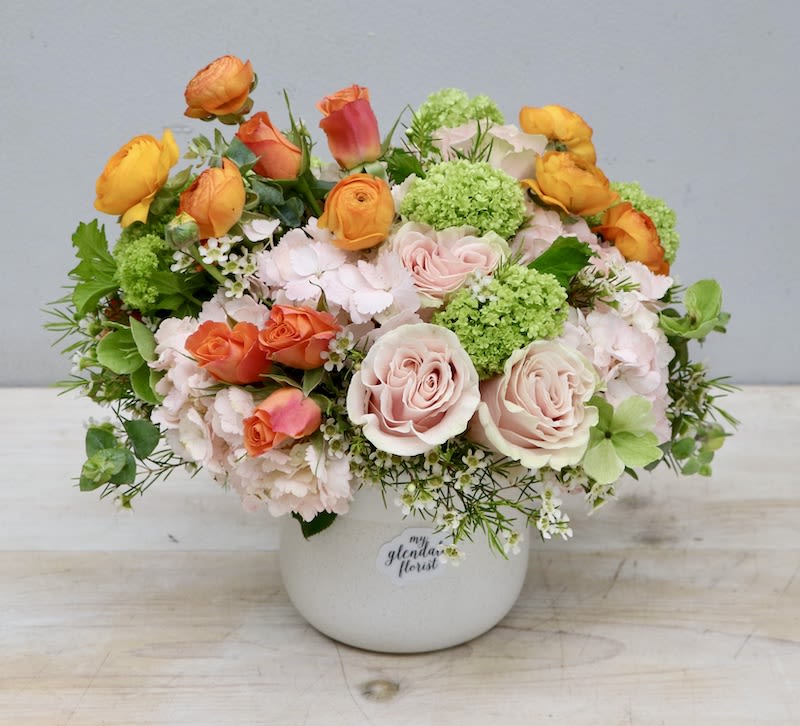 Low arrangement of peach roses, orange ranunculus, and green blooms in a white ceramic pot.