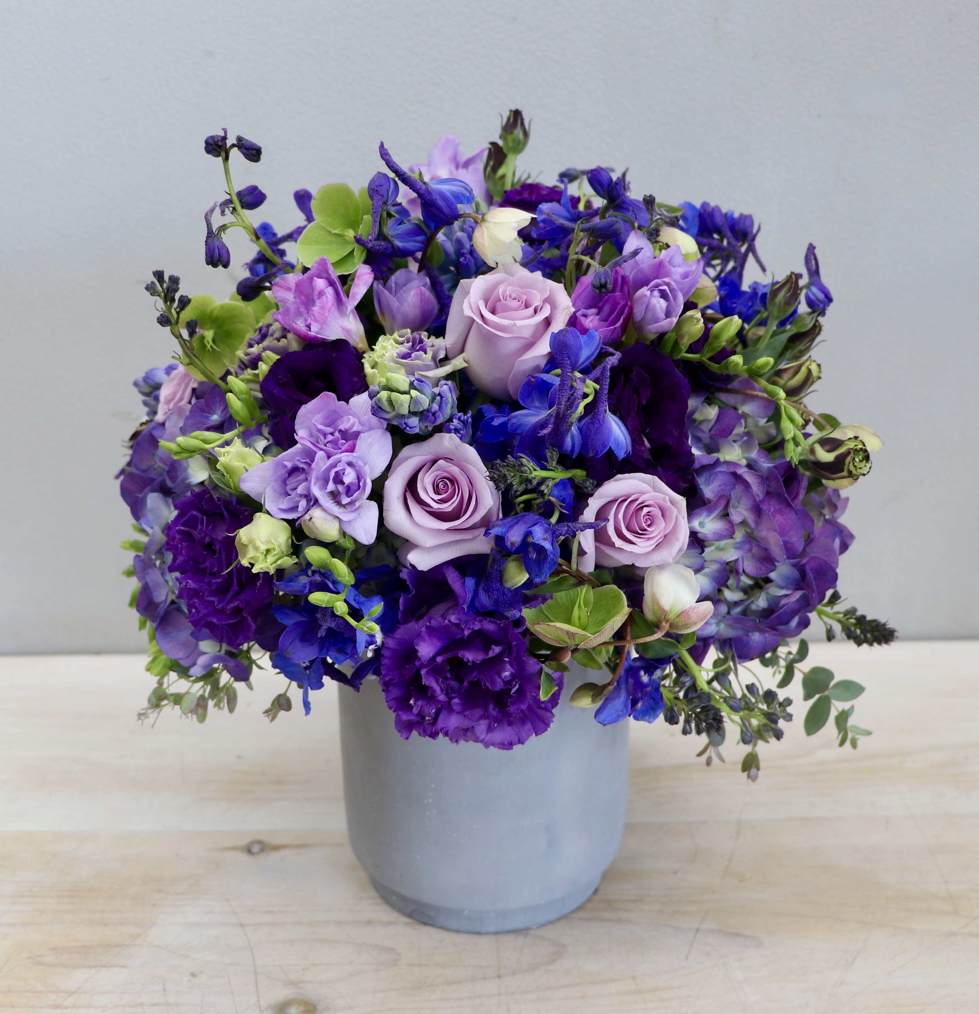 Round arrangement of purple, blue, and lavender flowers in a gray cylinder vase