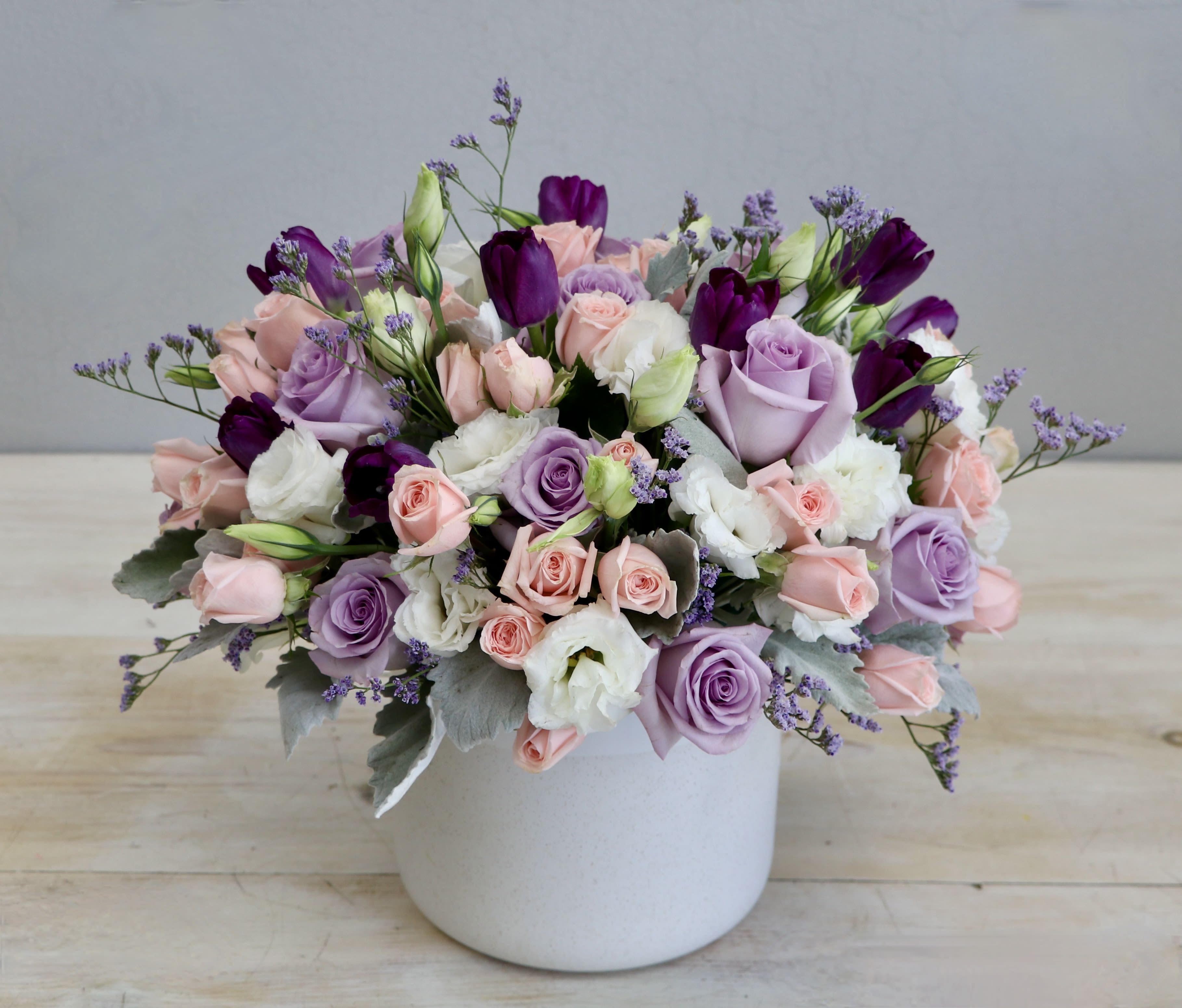 Low pastel arrangement of pink and lavender roses with white blooms in a white ceramic pot