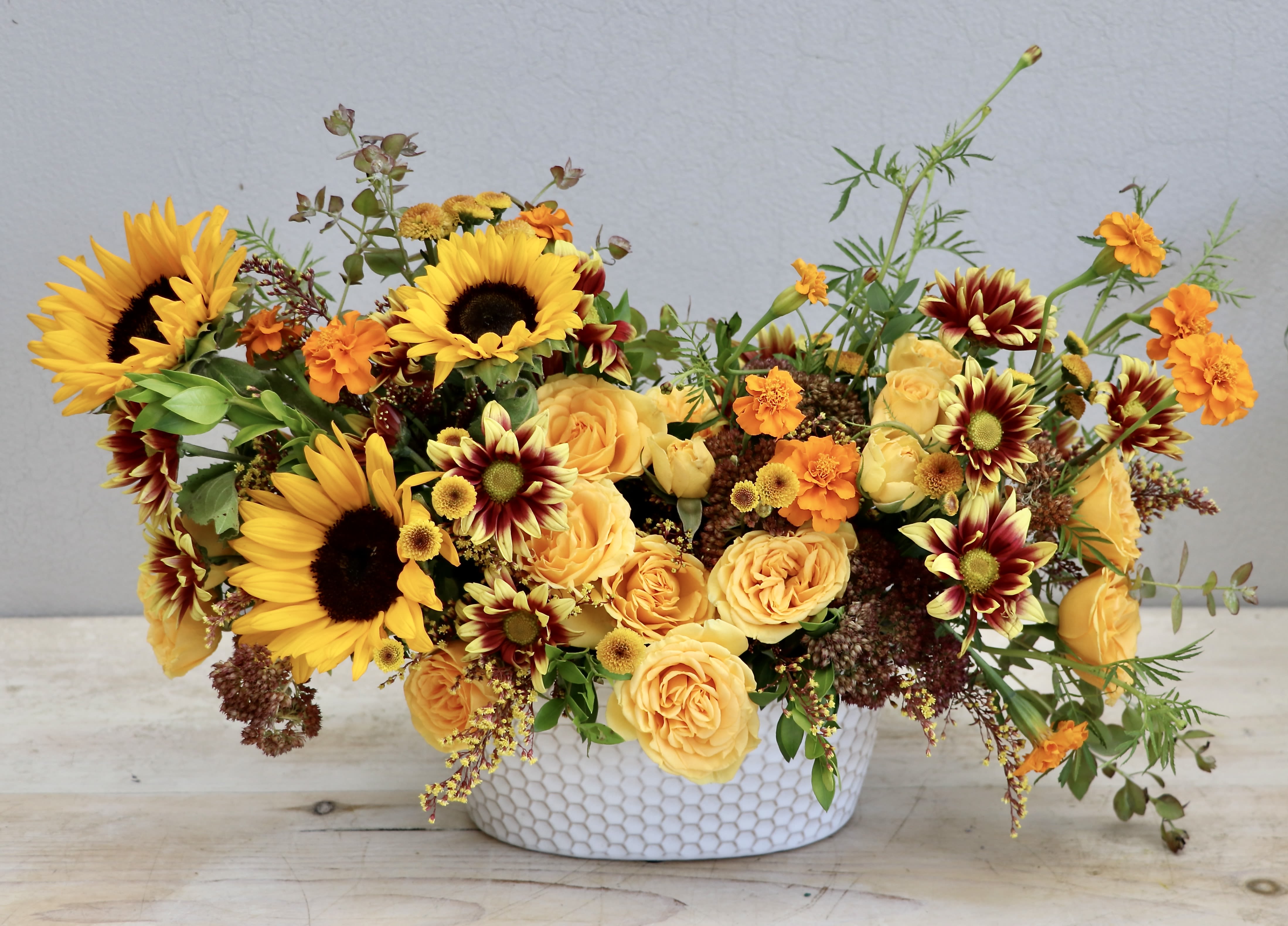 Low oval arrangement of yellow sunflowers, roses, and orange blooms in a white textured ceramic bowl