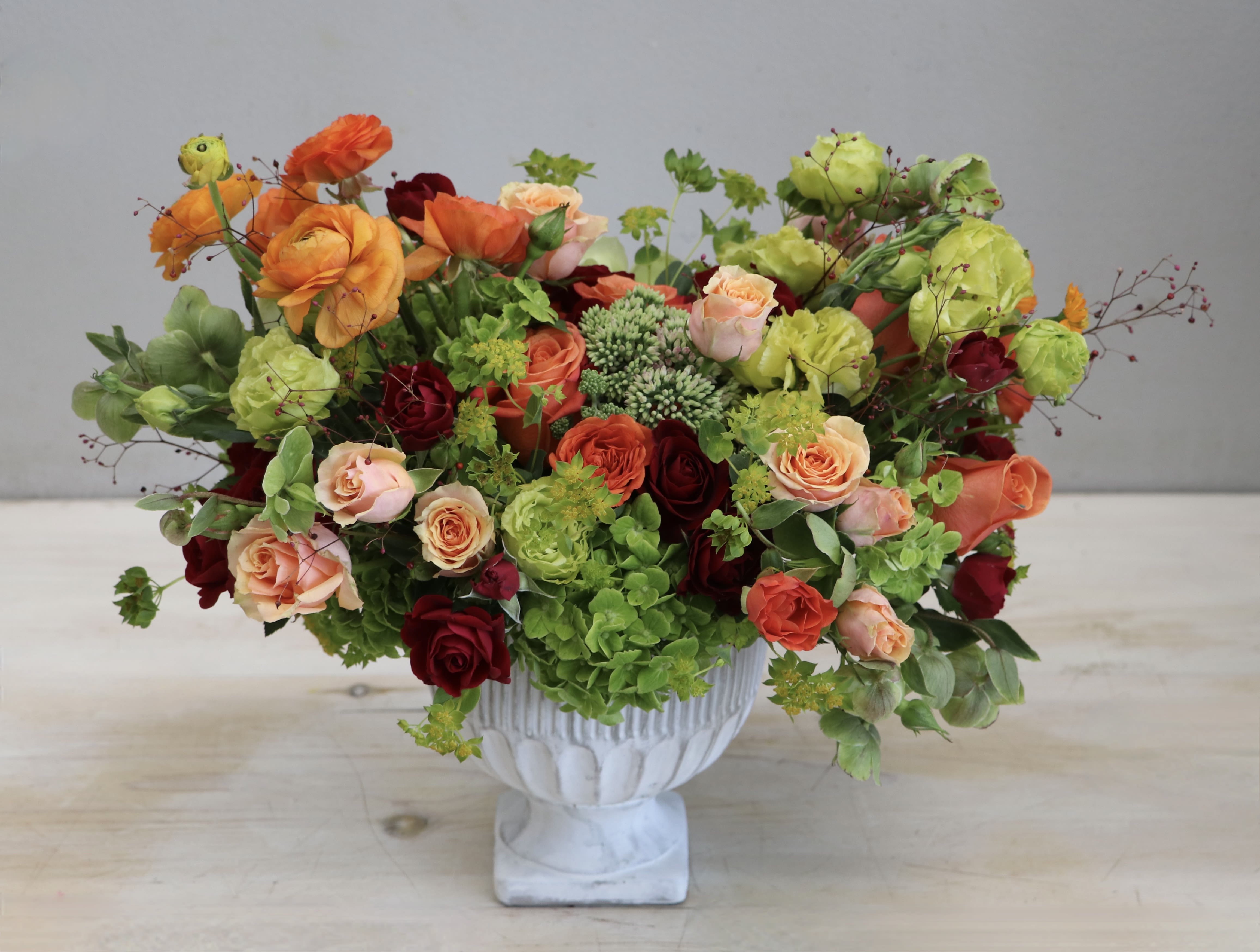Lush arrangement of orange, peach, red, and green flowers in a white pedestal vase