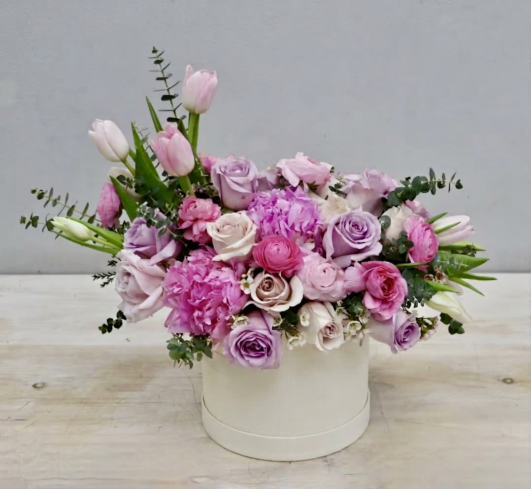 Sweet Lavender Peony Hat Box - My Glendale Florist - This hatbox is filled with lavender and blush roses, tulips, and fragrant pink peonies. A beautiful balance of pastels and bright pinks. This dreamy arrangement is a perfect gift for any celebration.   The standard size hatbox diameter is 7" and the arrangement is about 12" high. Each upgrade is filled with more blooms and a larger box.