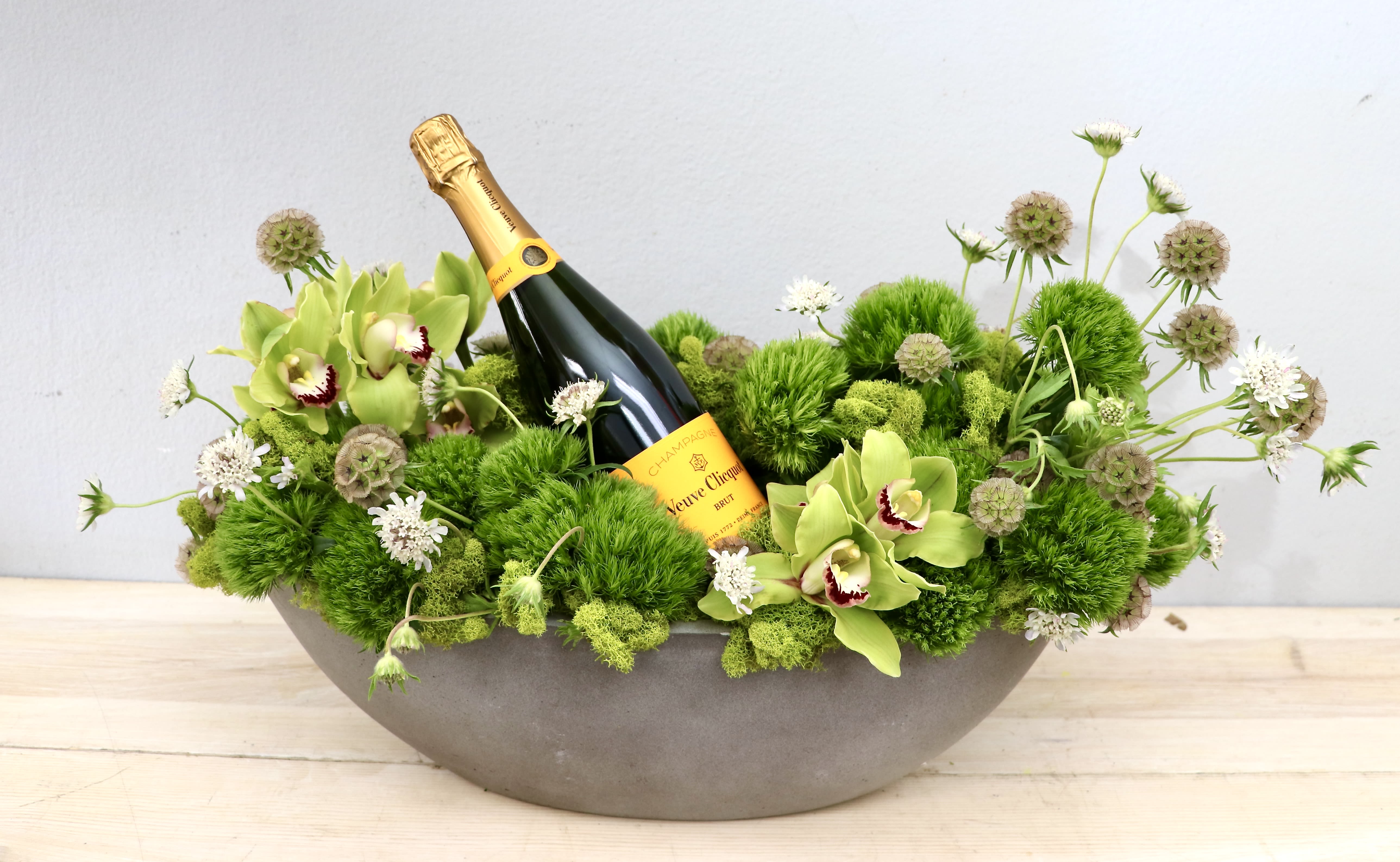 Gentlemen's Bottle - Glendale Flowers - Pale green orchids with dark centers tuck into a playful, low-profile celebration arrangement built around a chilled champagne bottle-an elegant flower arrangement perfect for a congratulatory gift or celebratory display. This Gentlemen's Bottle arrangement is ideal for anniversaries, promotions, housewarmings, or any occasion when you want the recipient to feel festively remembered.  A shallow gray bowl holds a dense, wide bed of bright green textured blooms and small white accents that spread outward around the bottle, creating lively contrast and pleasing horizontal lines. The pale green orchids provide a fresh focal point against the vivid greens and crisp whites, while the compact composition makes it easy to place on a buffet, coffee table, or office desk.  Give this to the person you're celebrating when you want them to feel noticed and honored-it's the kind of gift that announces congratulations without saying a word. Designed for immediate display, moderate care (keep water fresh and remove wilted blooms), and sized as a low centerpiece, it arrives ready to set and enjoy.