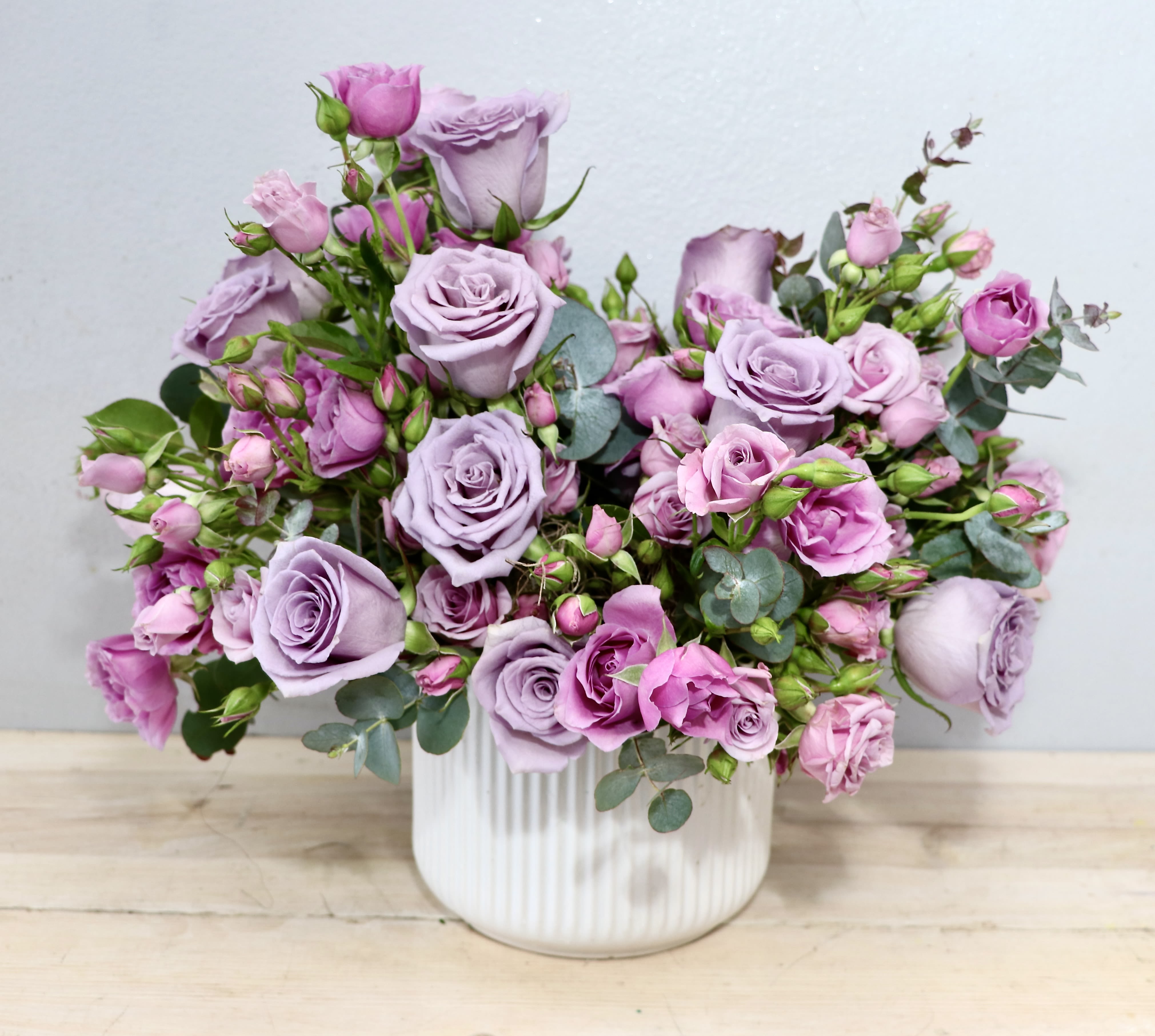 Garden Lavender - MGF - Lavender roses gathered in a low, garden-style cluster create a romantic birthday bouquet or anniversary gift that reads as effortlessly elegant. The pastel mix highlights and green eucalyptus gives the recipient a gentle, calming impression-perfect for celebrating a milestone or simply making someone feel truly cherished.  The composition is broad and abundant rather than tall, with roses in varying stages of bloom to add texture and a natural, just-picked look.   This is the kind of floral gift that makes the person you're thinking of feel seen and celebrated-suitable for anniversaries, birthdays, springtime congratulations, or a thoughtful "just because." Arrives ready to display in the included ceramic vase; keep water fresh and trim stems occasionally to extend bloom life.