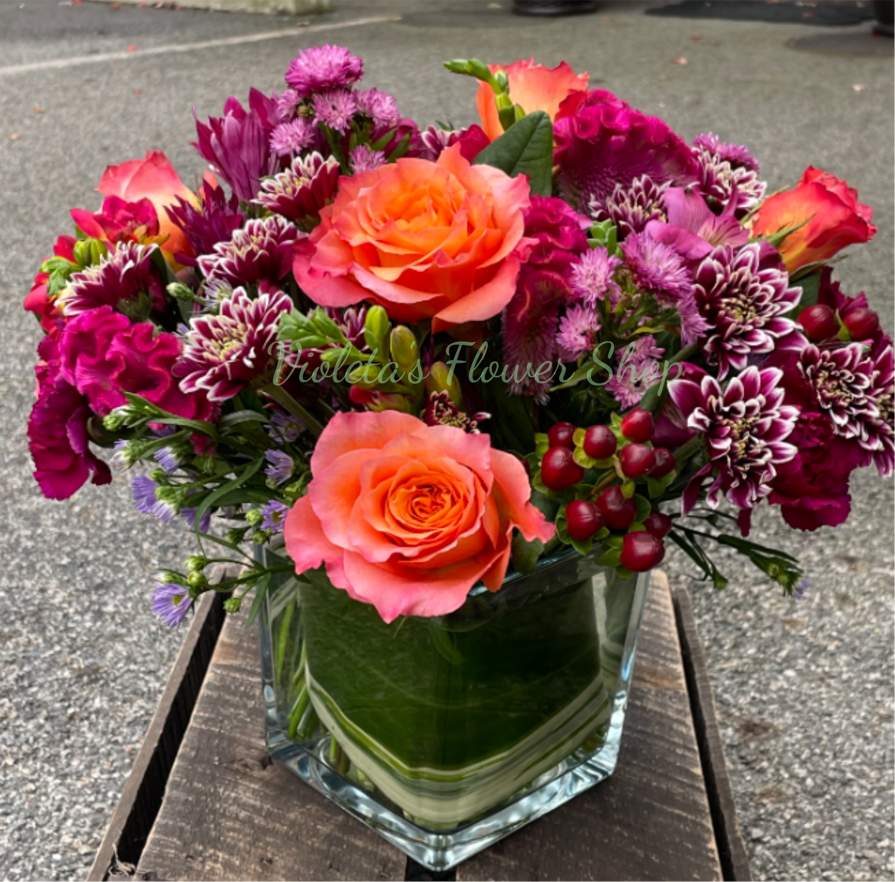 Sunsets With You Arrangement - Vibrant orange roses take center stage in this mixed flower arrangement, making it an immediate eye-catcher for a birthday bouquet, thank-you gift, or everyday celebration. Sunsets With You Arrangement pairs those warm focal roses with magenta, purple, pink, red and white blooms to recreate the glow of a summer dusk-she'll feel noticed and celebrated the moment the vase is set down.  The arrangement is full and rounded, packed with roses, chrysanthemums, carnations, asters and small daisy-like filler flowers that add ruffled texture and visual depth. It arrives in a clear square glass vase so stems and water are visible, lending a freshly hand-gathered look that suits modern tabletops or a festive centerpiece. Colors are saturated-bright oranges and warm pinks balanced by cool purples and whites-for a lively, layered composition that stands about the height of the vase plus the blooms.  