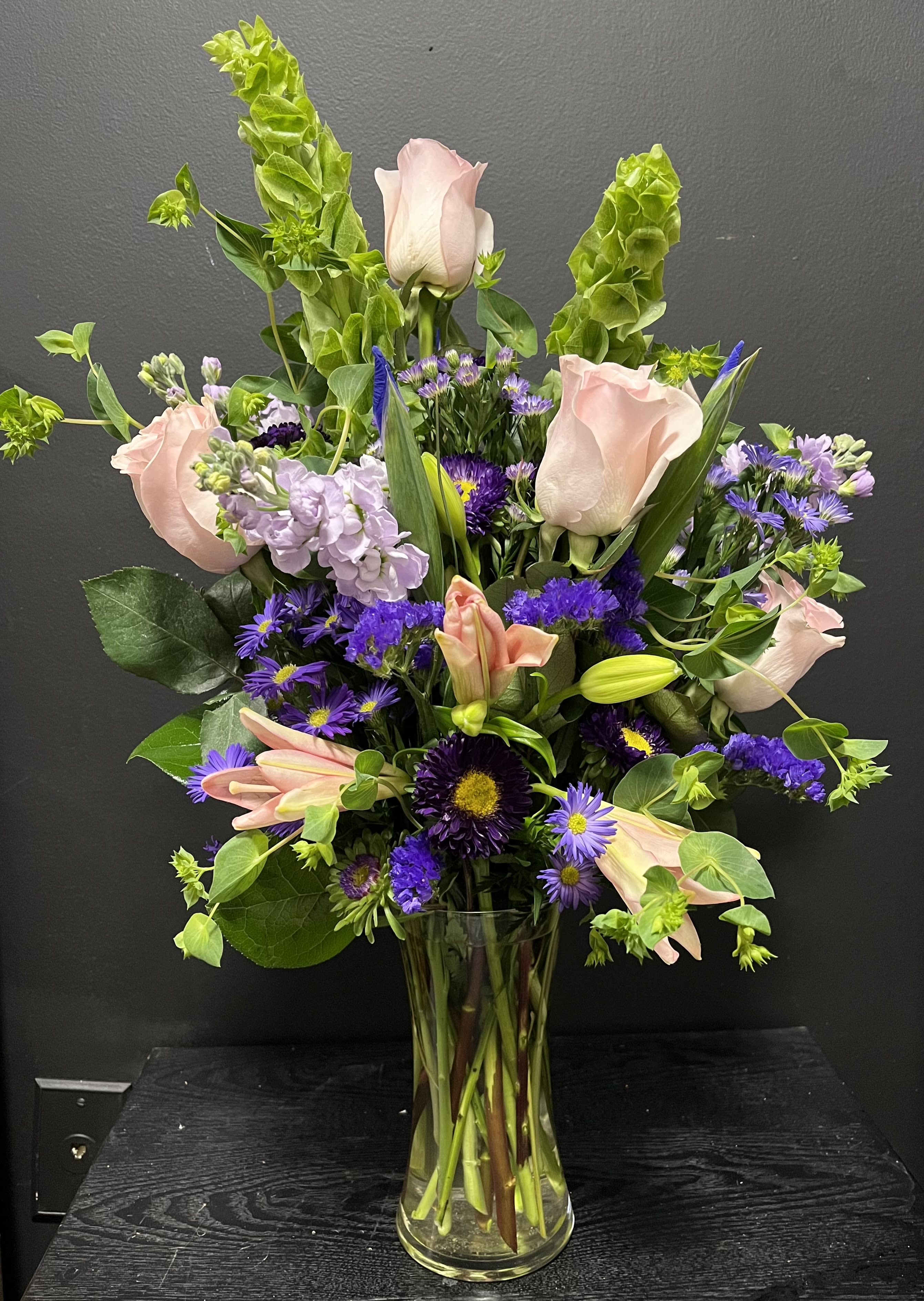 Tall vase arrangement of pink roses and lilies with purple flowers and green spikes.