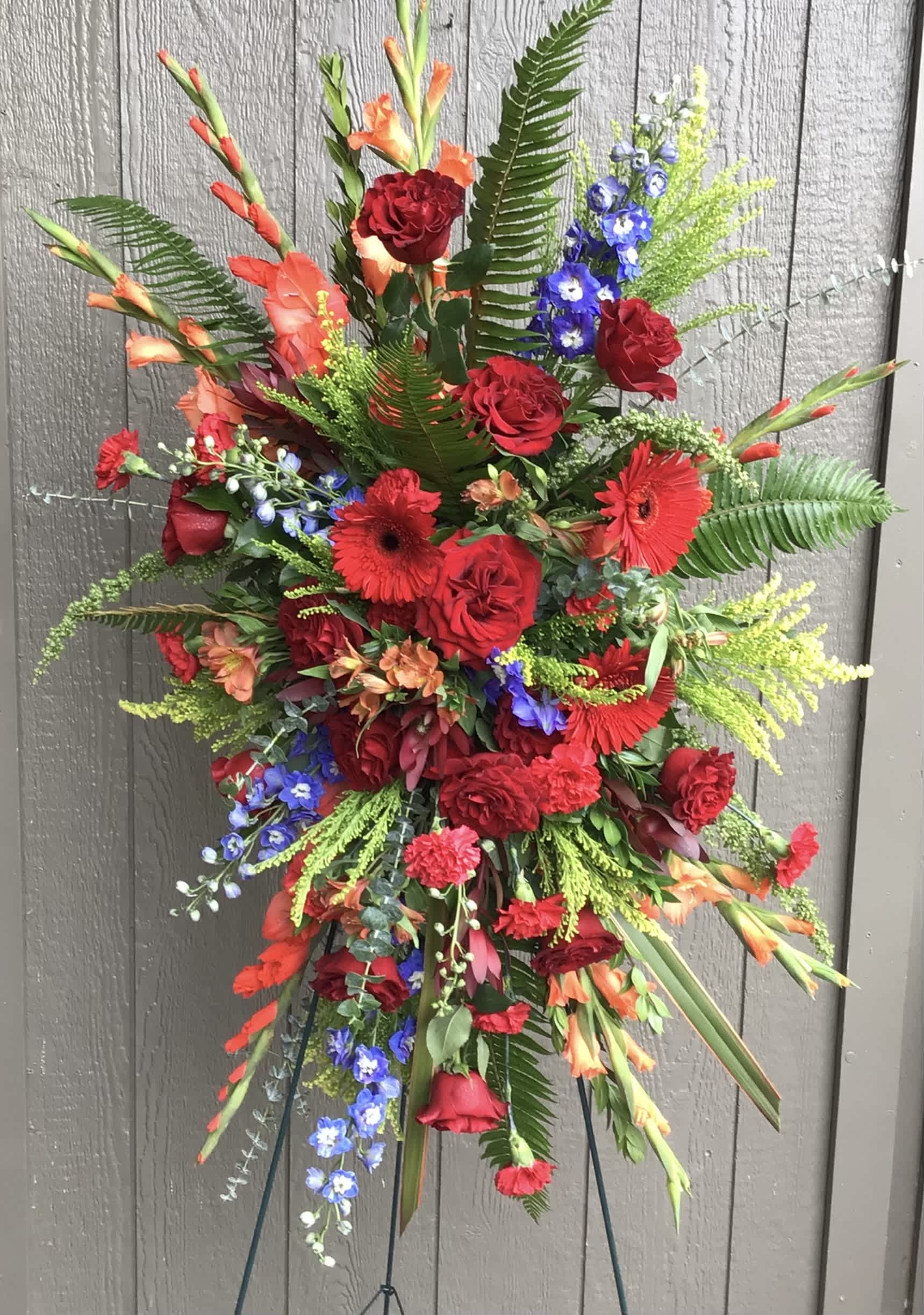 Tall standing floral spray with red roses, gerbera daisies, and blue accents