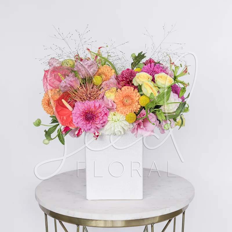 Bright mixed arrangement of pink, orange, and yellow flowers in a white square vase.