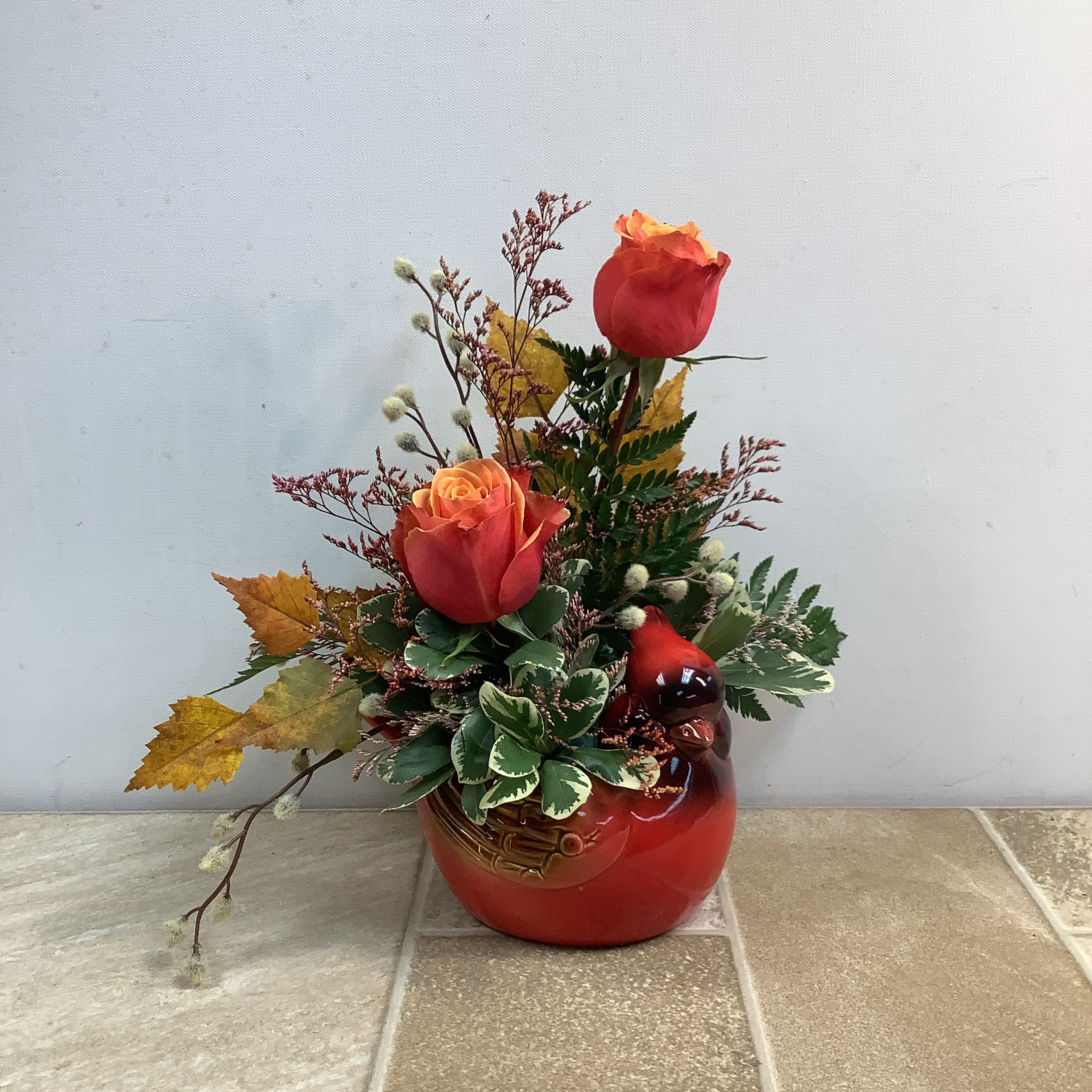 Two red-orange roses in a red ceramic vase with autumn leaves