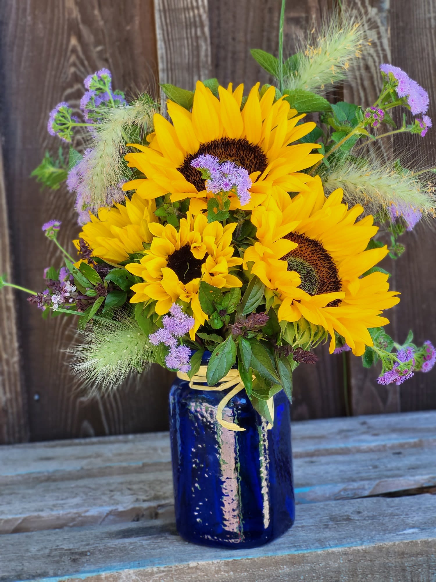 Feels Like Summer - A cobalt blue vase filled with locally grown sunflowers, ageratum and bunny tails! It's a jar full of sunshine and smiles!