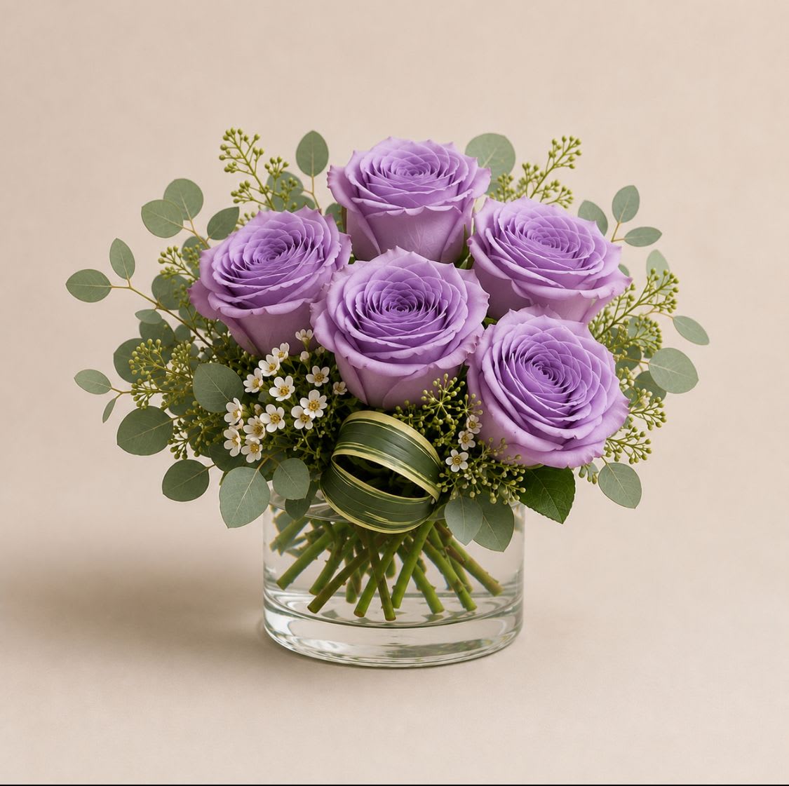 Twilight - A compact dome of five lavender roses creates a calm, romantic focal point for a birthday bouquet, anniversary roses gift, or a thoughtful thank-you arrangement. The soft purple tones paired with green and white accents read as quietly elegant-perfect for the person you want to make feel seen without fuss.  Clusters of airy eucalyptus and small white filler flowers surround the roses for texture and a fresh scent, while a clear glass vase keeps the silhouette light and modern. A green decorative ribbon finishes the look, making it ready to display on a bedside table or office desk; size is a neat tabletop arrangement ideal for small spaces and easy care-fresh water and a clean vase help the roses last longer.