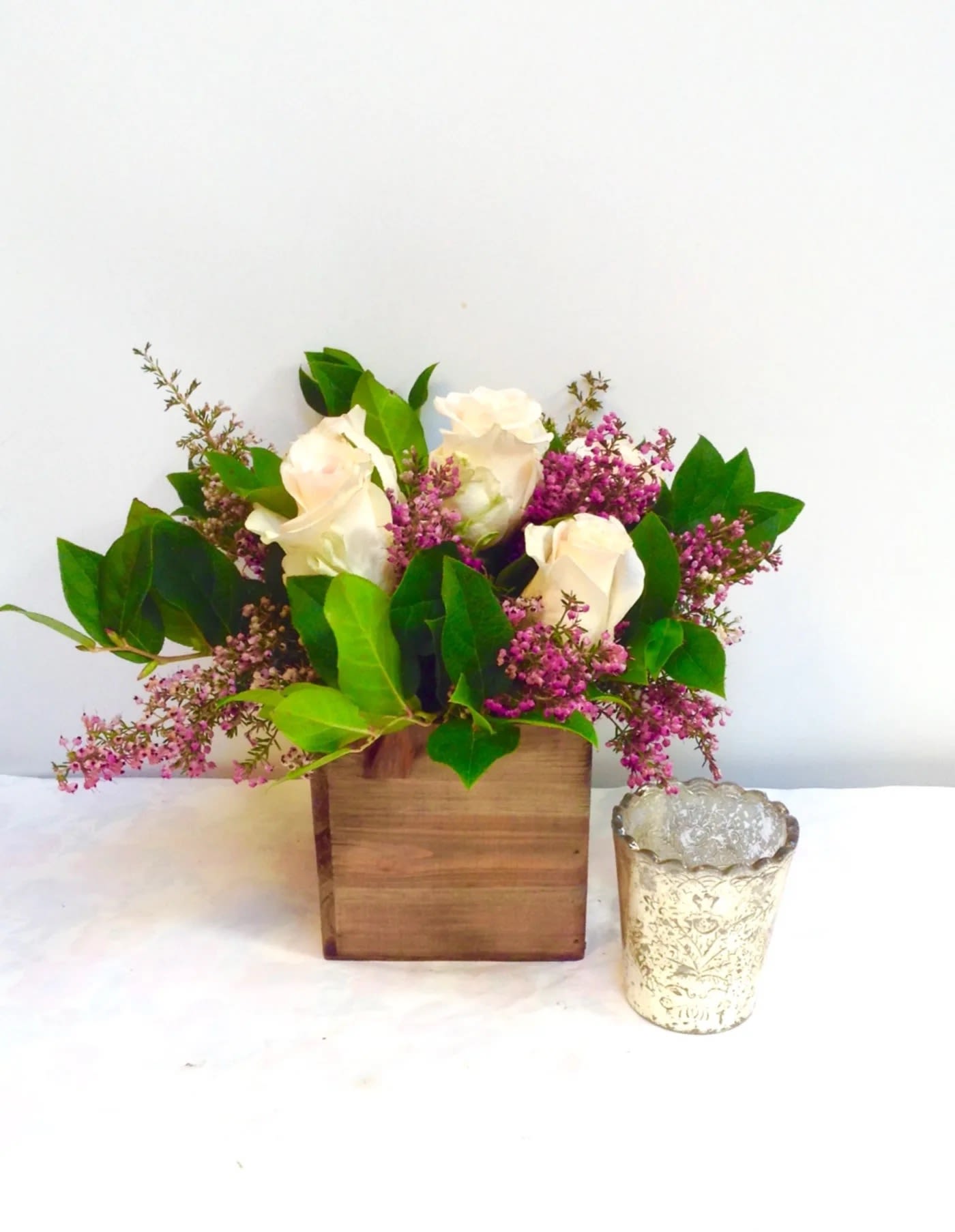 White roses and pink filler flowers in a wooden box with a glass votive beside it