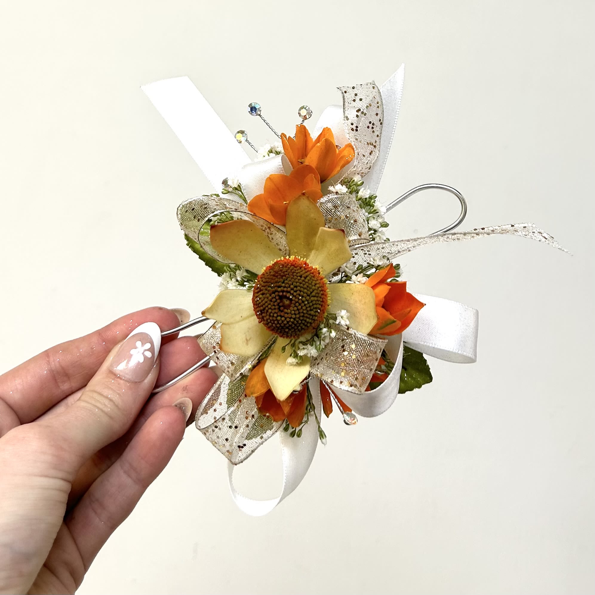 Orange Wrist Corsage - A bright orange wrist corsage that feels both celebratory and polished - perfect as a prom corsage or for graduation and other formal events. The warm orange blooms surround a central yellow-green flower head that draws the eye, while white satin and sheer glitter ribbon add a soft, dressed-up contrast that catches the light.  This wristlet features decorative wire accents, rhinestone-topped picks for sparkle. The compact scale keeps it comfortable on the wrist without overwhelming a dress or suit; colors include orange, white, yellow and mixed tones for a lively, autumn-tinged palette that works year-round.  Made to complement formal attire and mark special moments, this corsage sends a cheerful, celebratory message to the person you're honoring. If you have flower preferences, include them in the special instructions and we'll do our best to accommodate.