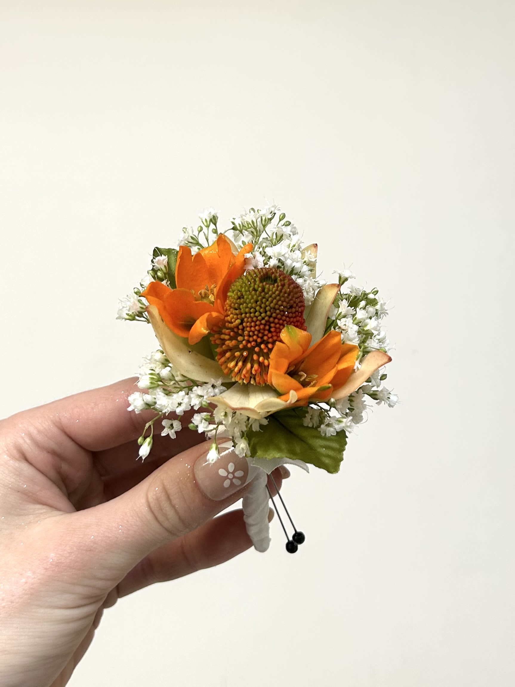 Orange Boutonniere - Bright orange blooms clustered around a textured central bloom give this boutonniere a bold, celebratory look perfect for prom or a wedding. The compact orange-and-white contrast - with tiny white filler flowers and pale petal accents - creates a fresh, lively impression that will make the person wearing it feel noticed and festive.  Hand-wrapped in white boutonniere wrap and secured with small black floral pins, the stemwork reads formal and handcrafted. Sized to sit neatly on a lapel, the arrangement includes boutonniere wrap and floral pins; if you have a flower preference, please note it in special instructions and we'll try to accommodate. This piece is designed to coordinate with orange, white, and green palettes and to hold up through an evening of photos and celebration.