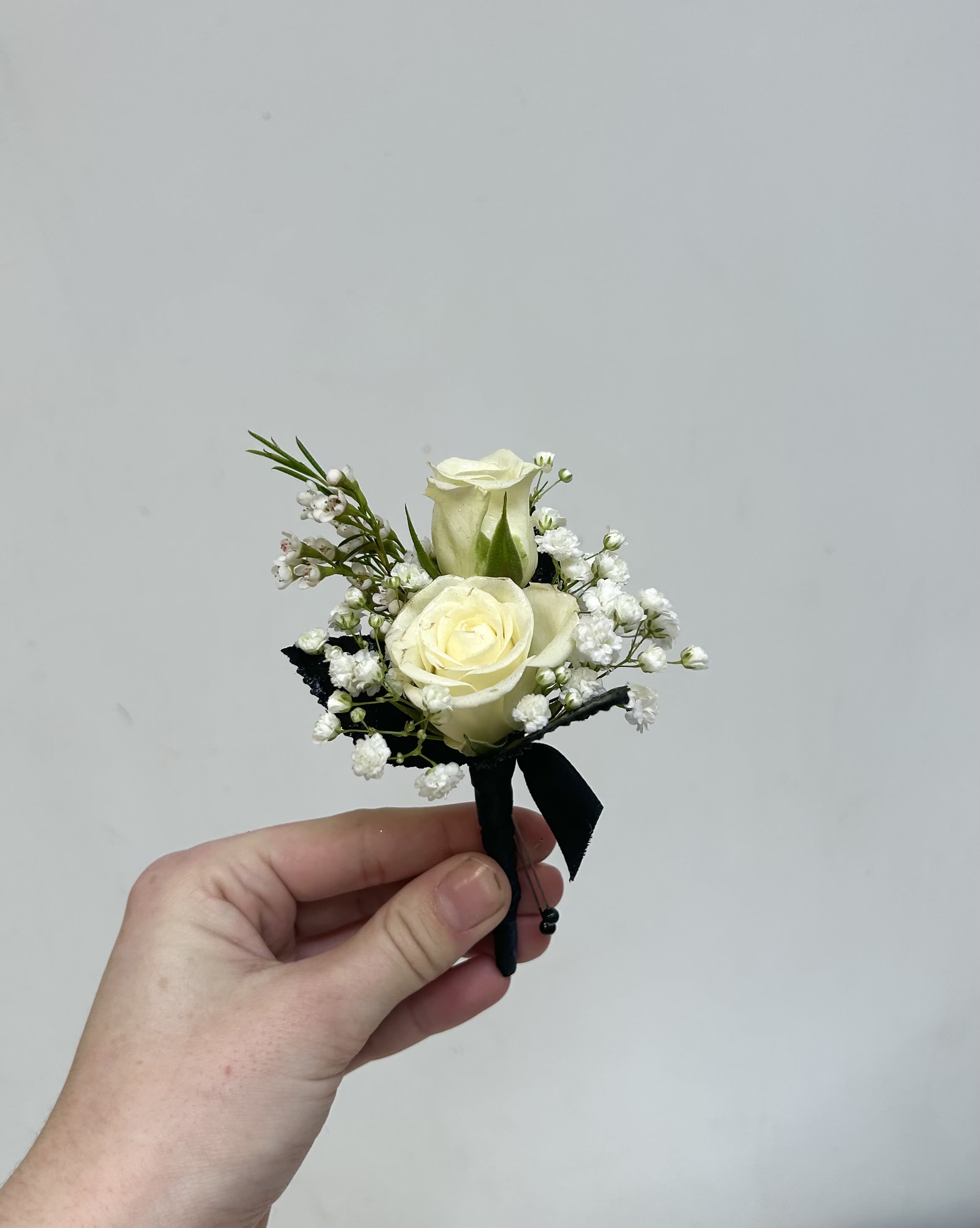 Black &amp; White Mixed Boutonniere - Two ivory-white roses sit close together in this compact boutonniere, surrounded by delicate white filler blooms for a refined, textured look-perfect for a prom or any formal event where classic black-and-white style matters. The contrast of the creamy blooms against a neat black ribbon gives a clean, elegant impression that will make the person wearing it feel polished and noticed without overpowering their outfit.  Hand-tied and finished with a boutonniere pin, this wearable floral accessory is small and lightweight for comfortable all-evening wear. The palette of white, green foliage, and black ribbon pairs easily with tuxedos or dark suits; if you have a flower preference, note it in the special instructions and we'll do our best to accommodate. For best results, keep cool and dry until pinned on-ideal for gifting the person you want to look especially put-together.