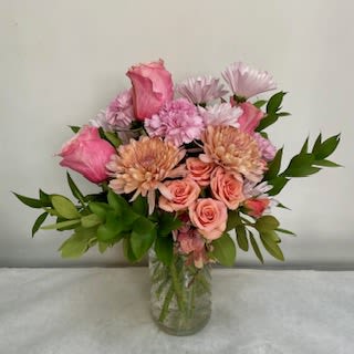 The Taylor - Soft, garden-fresh colors and a relaxed round shape make this flower arrangement a gentle way to celebrate someone special-perfect as a birthday bouquet, anniversary surprise, Mother's Day gift, or a thoughtful "thinking of you" pick-me-up. The pink roses and peach chrysanthemums create a warm, affectionate palette while pale daisy-like blooms and broad green foliage add lightness and a casual, hand-gathered feel that will make the recipient feel seen and comforted.  This arrangement is built in a clear glass vase that lets the stems show through and complements the soft pinks, peaches, greens, and whites. The mix includes pink roses, peach-toned chrysanthemums, and delicate white-lavender daisy-style flowers, arranged in a loose, rounded garden-style composition rather than a tightly formal bouquet. The overall size and fullness make it an easy centerpiece for a kitchen table or a bright addition to a bedside or office desk.