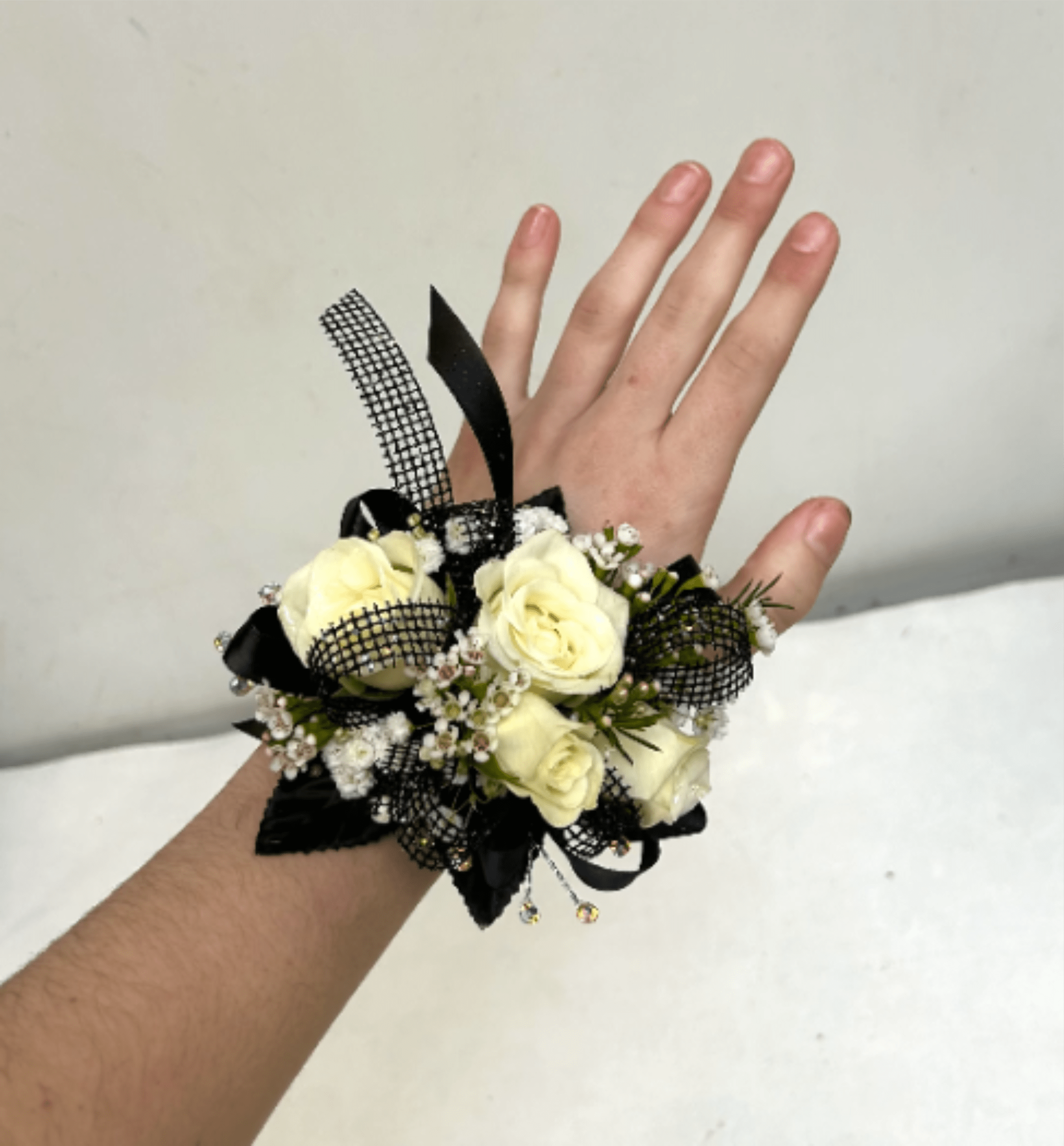 Black &amp; White Mixed Wrist Corsage - Cream-white roses gathered in a compact wrist corsage create an elegant, modern statement perfect for prom or other formal occasions. The monochrome palette-soft white blooms offset by black ribbon loops, black mesh accents, and sparkling rhinestone details-gives the recipient a refined, slightly dramatic look that reads timeless whether it's worn to a dance, wedding, or gallery opening.  This wrist corsage features several tightly clustered cream-white roses with small white filler blooms and greenery for texture, all secured to a comfortable wrist band. Black satin ribbon and black mesh ribbon weave through the arrangement for contrast, while rhinestone accents add a subtle touch of sparkle. The compact size keeps the design graceful and wearable without overwhelming the wrist.  Choose this corsage to make someone feel celebrated and elegantly seen on their special night; it pairs well with black, white, or jewel-toned attire. If you have a flower preference, note it in special instructions and we will try our best to accommodate. Care: keep cool and mist lightly until wear.