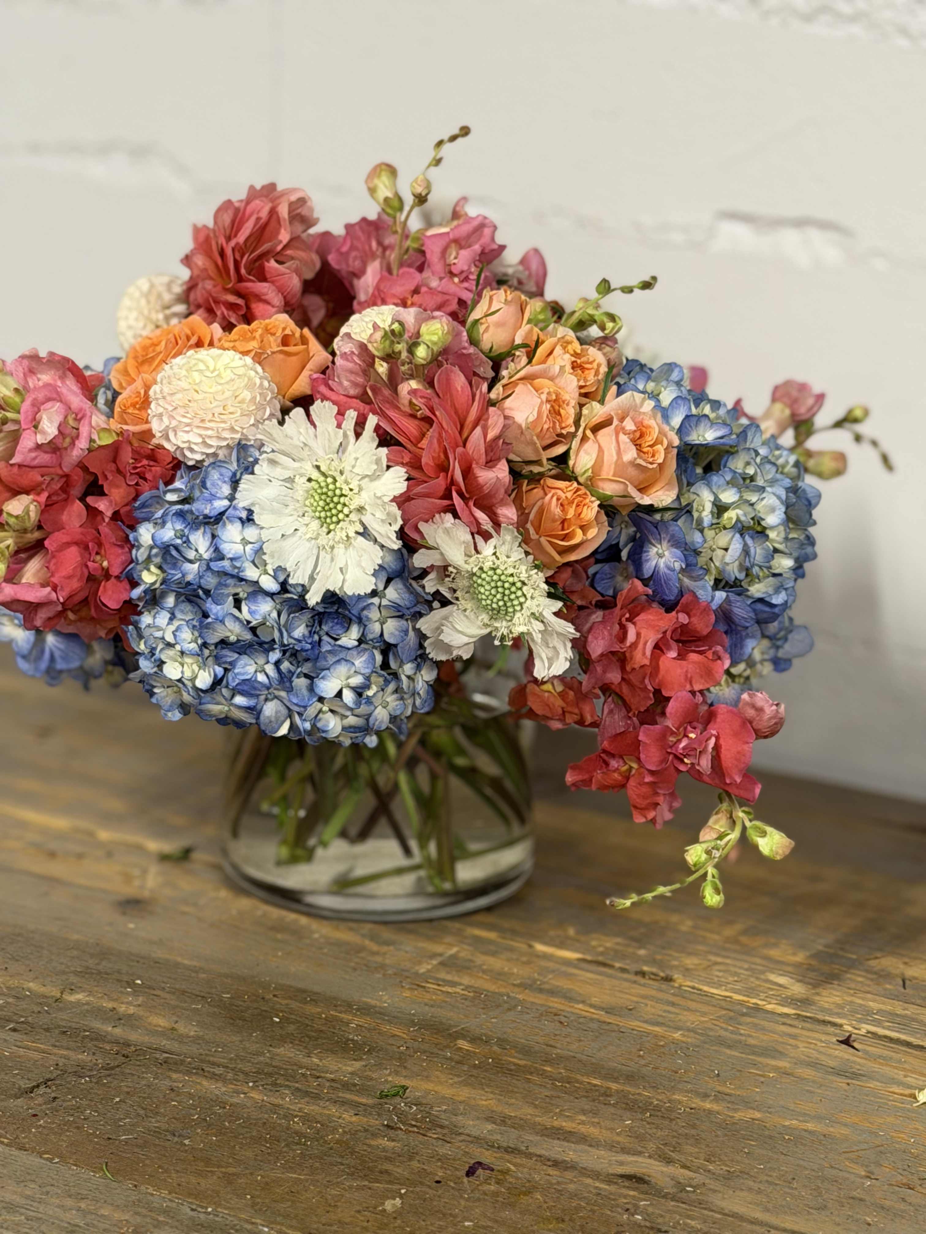 Mixed bouquet of blue hydrangeas, peach roses, and pink flowers in a glass vase