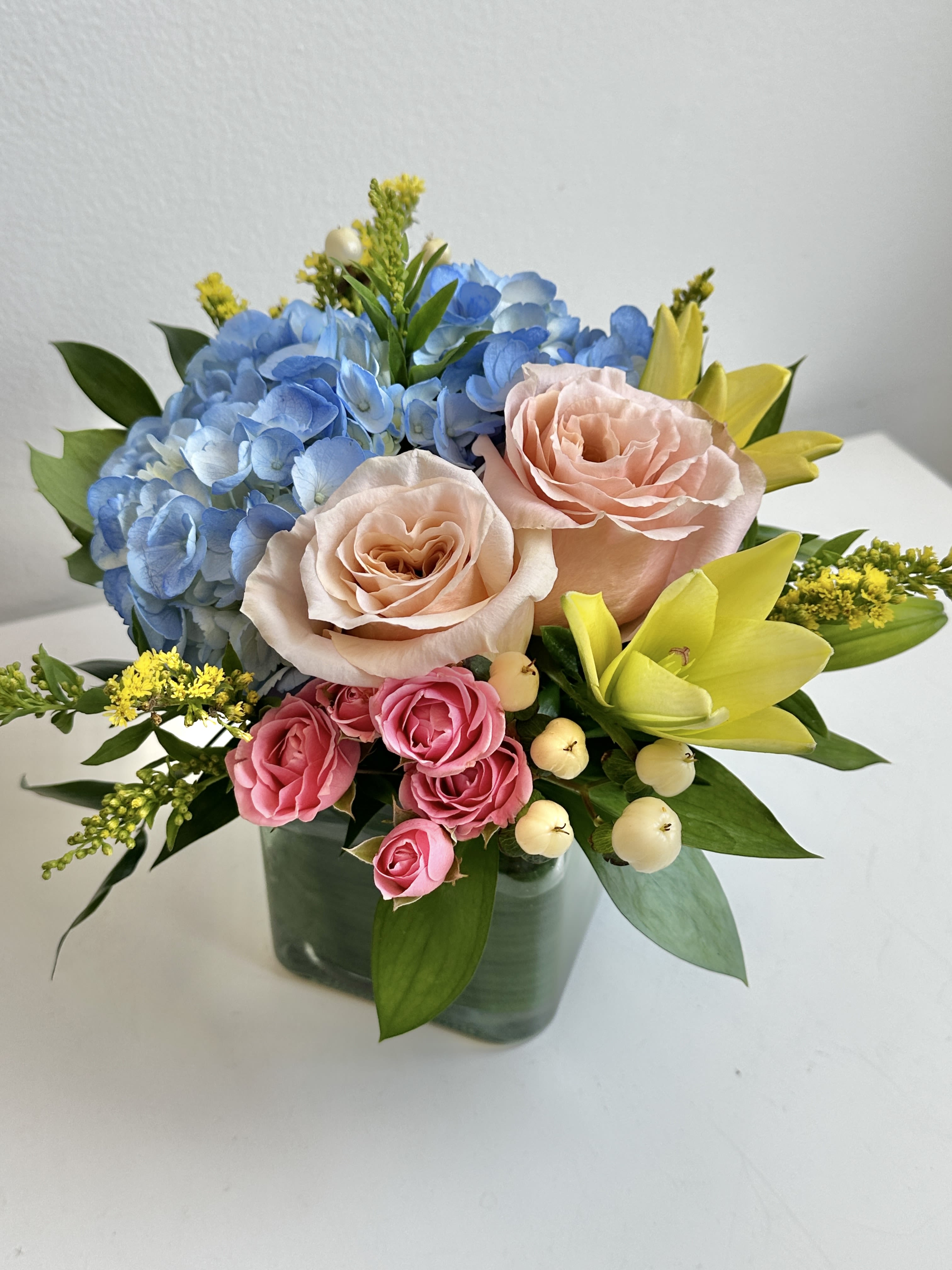 Bouquet of blue hydrangeas, peach roses, pink spray roses, and yellow lilies in a glass vase