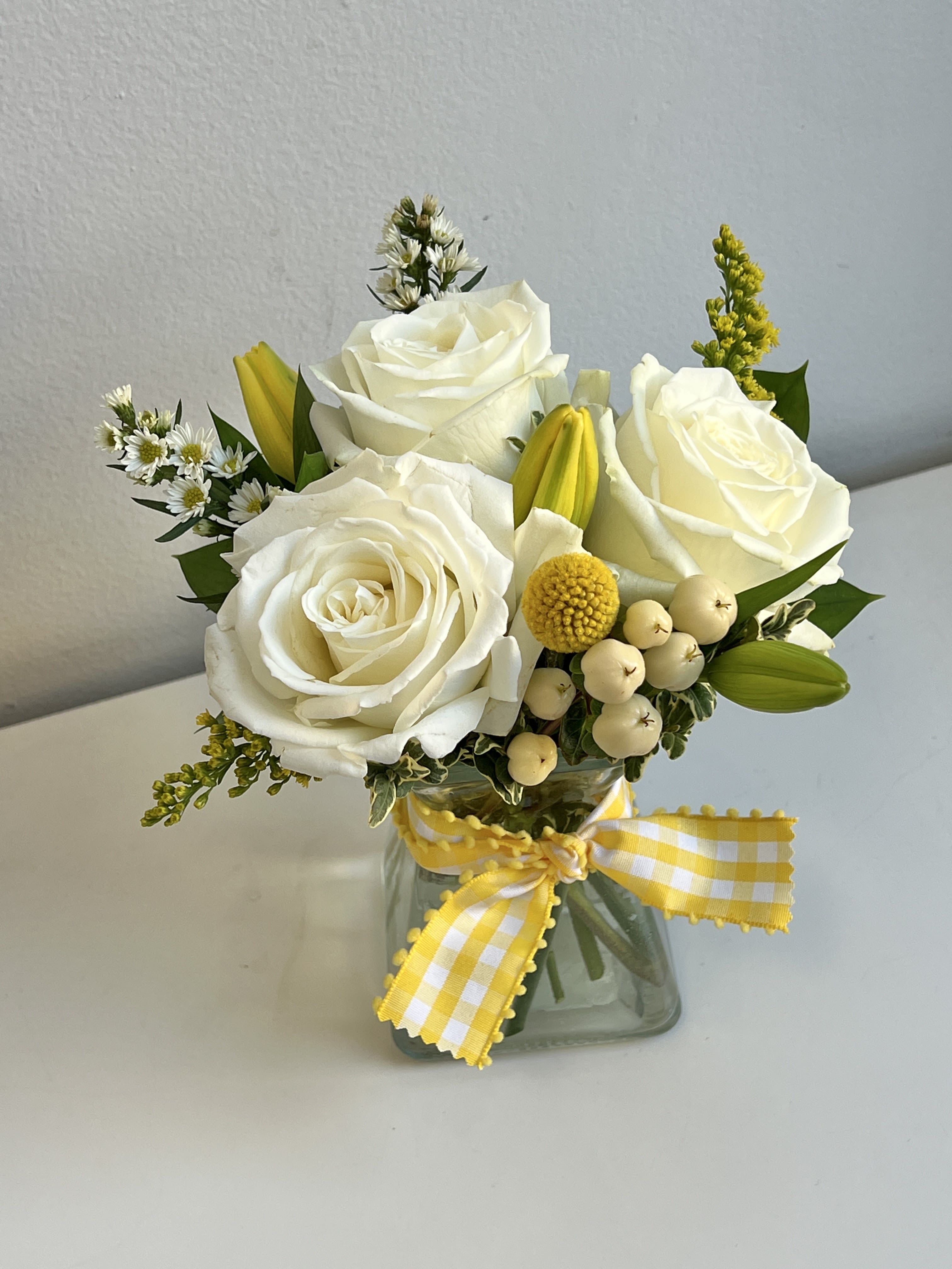 White roses and yellow accents in a glass vase with a gingham ribbon
