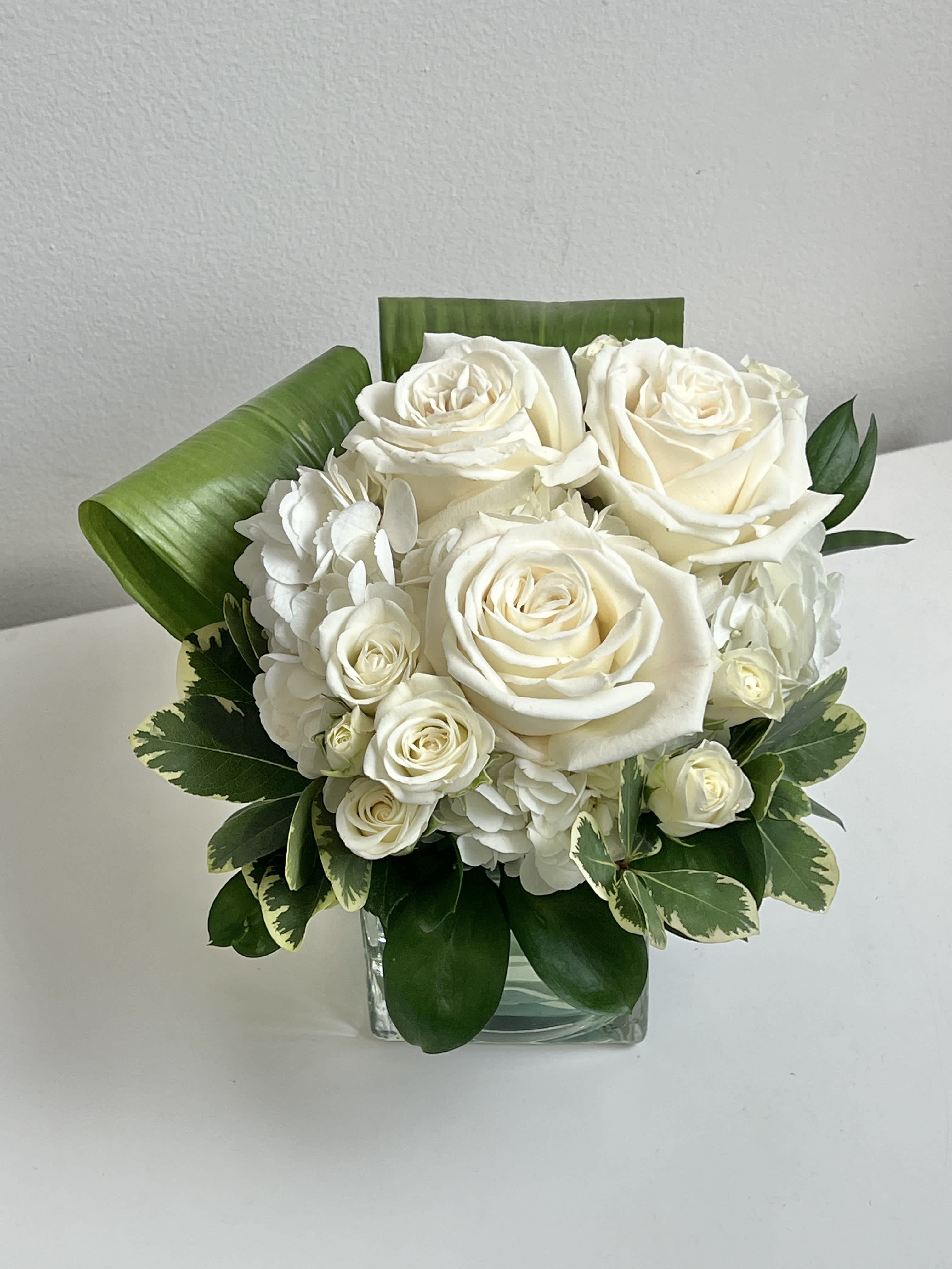White rose bouquet in a clear square vase with green foliage